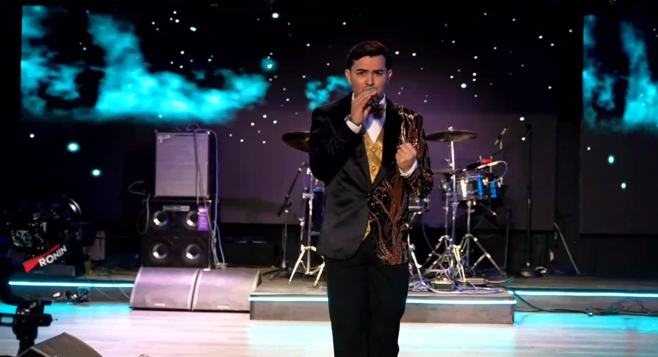A man in a black suit with intricate gold and brown embroidery, holding a microphone and singing on stage, with a background of a starry sky and blue nebula visuals, and musical instruments behind him. La voz Guacalito, Alberto Jimenez cantante latin