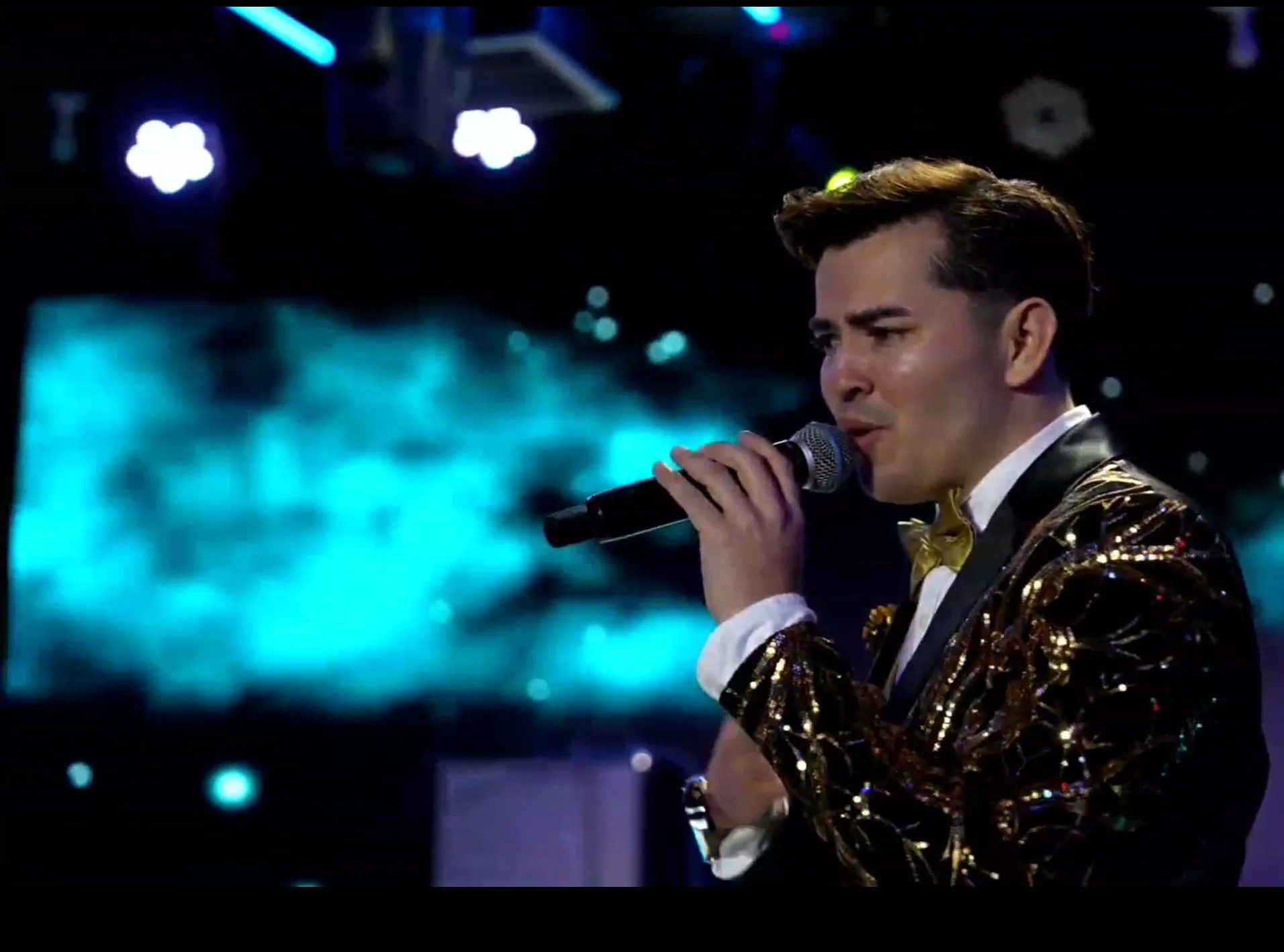 A man singing into a microphone on stage, wearing a shiny gold and black jacket with a white shirt and a gold bow tie, with a blurred digital screen background. La voz Guacalito, Alberto Jimenez cantante latino