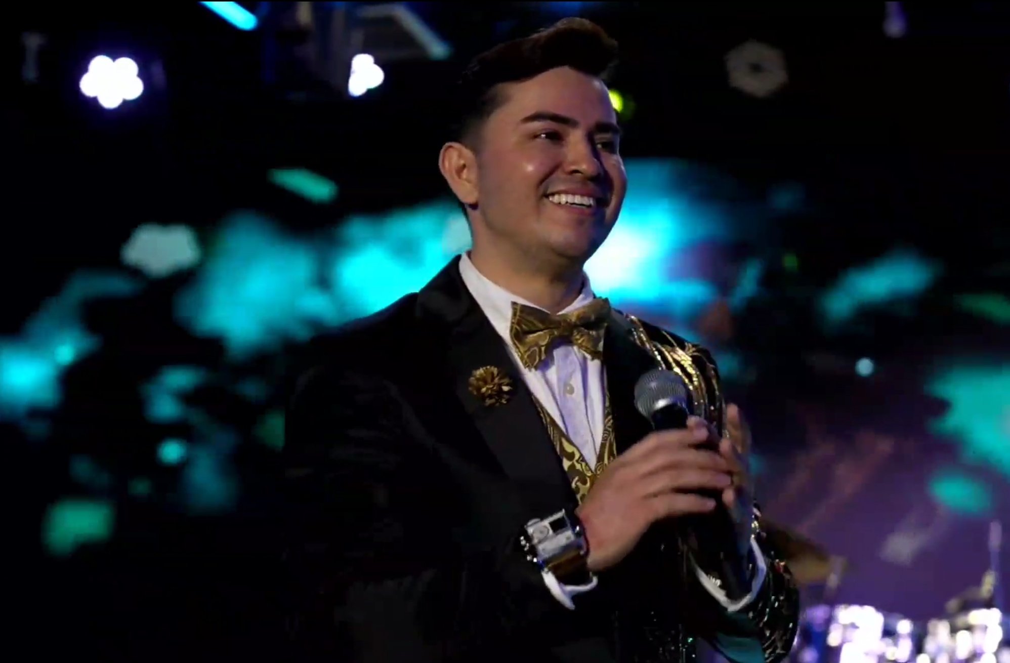 A man in a black tuxedo with gold accents and a bow tie, smiling and holding a microphone, stands on a colorful stage with bright blue and purple lights in the background. La voz Guacalito, Alberto Jimenez cantante latino