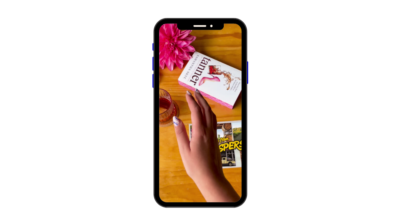 A smartphone with a hand touching a photo on the screen, surrounded by a pink flower and a glass of water, on a wooden table with a magazine and images.