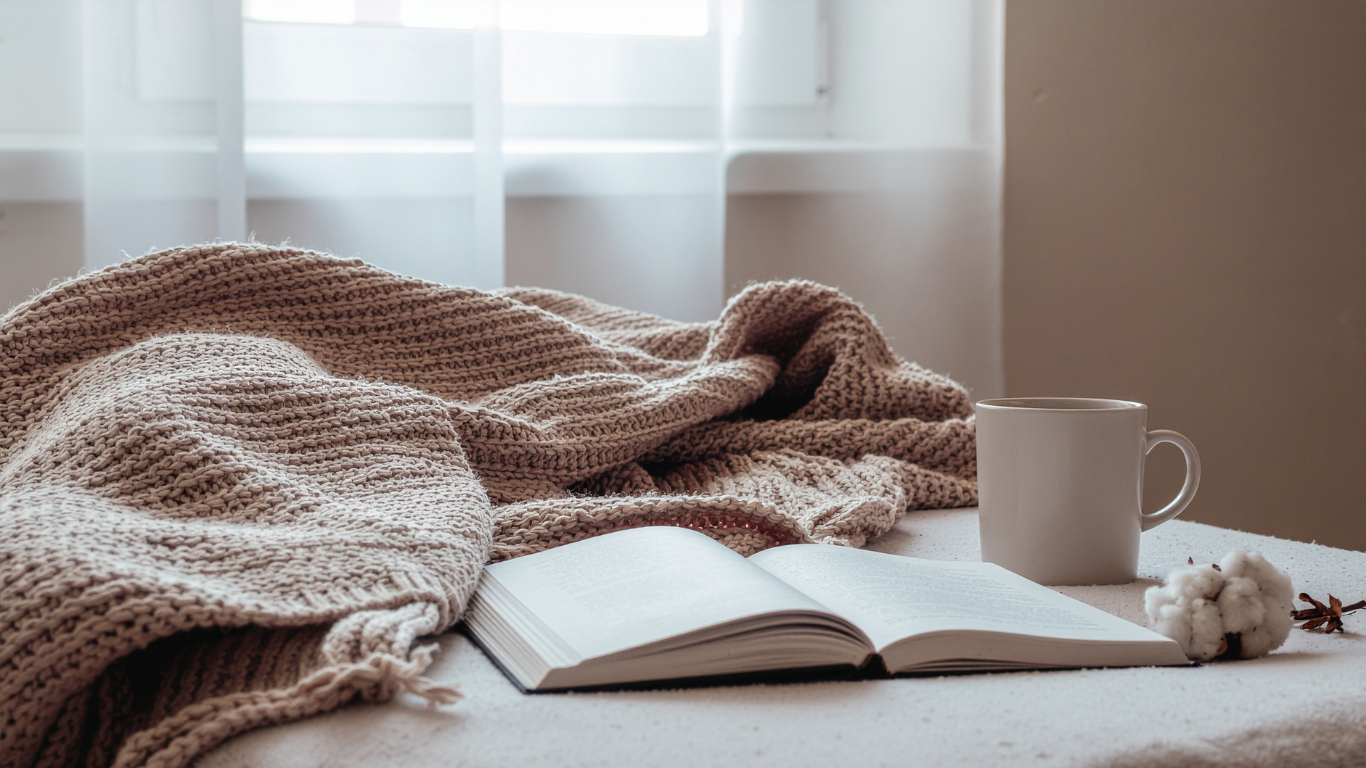 A cozy bed with a knitted blanket, an open book, a white mug, a cotton flower, and a sprig of cinnamon on a white surface near a window with sheer curtains.
