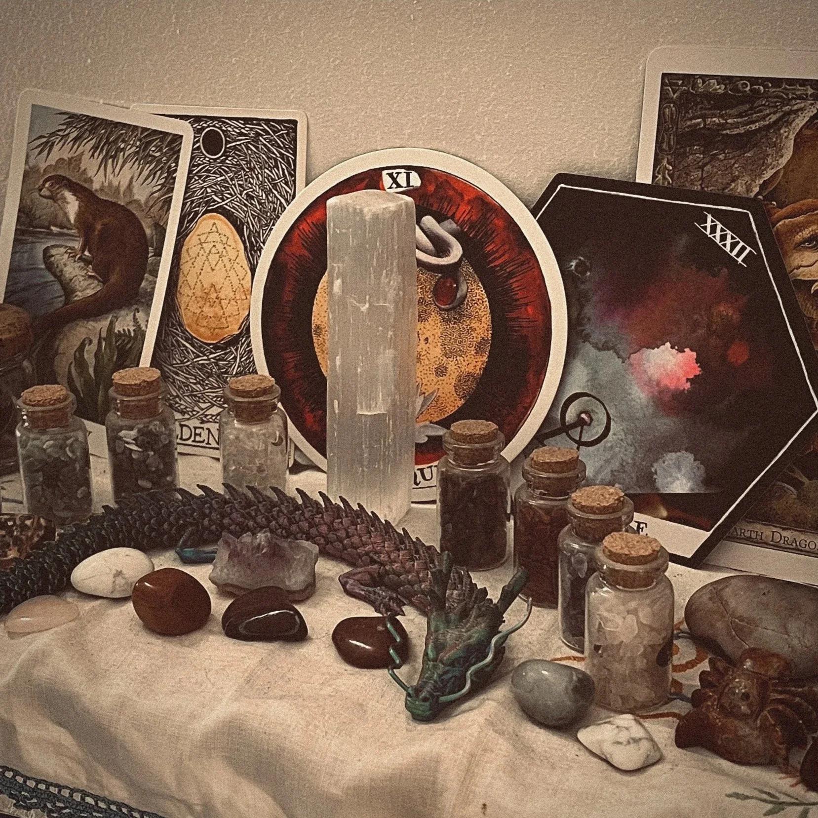 A display of various crystals, stones, and minerals, including small glass bottles with cork stoppers, some with dark crystals or powders inside. There is a carved dragon figurine with green and black accents at the front. Behind the display, tarot cards and a circular piece of artwork with a red and black theme are visible.