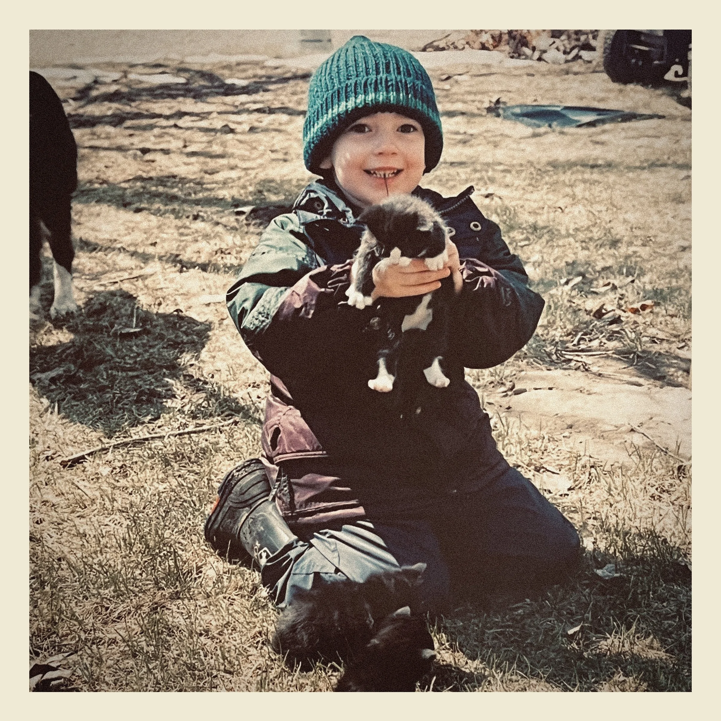 A young child wearing a blue and black striped knit hat, a camouflage jacket, and gloves, smiling and holding a small puppy with black and white fur outdoors on autumn leaves.
