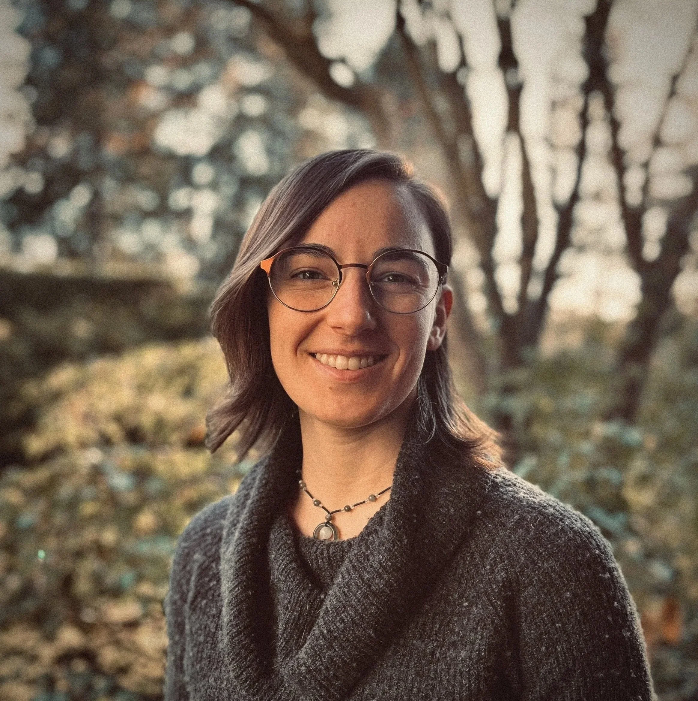 Partial view of a smiling woman with glasses and brown hair, standing outdoors with blurred trees and leaves in the background.