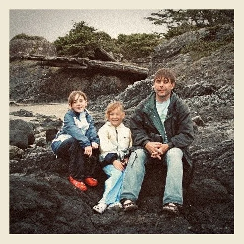 Three children sitting on rocks by a river, surrounded by trees and large boulders.