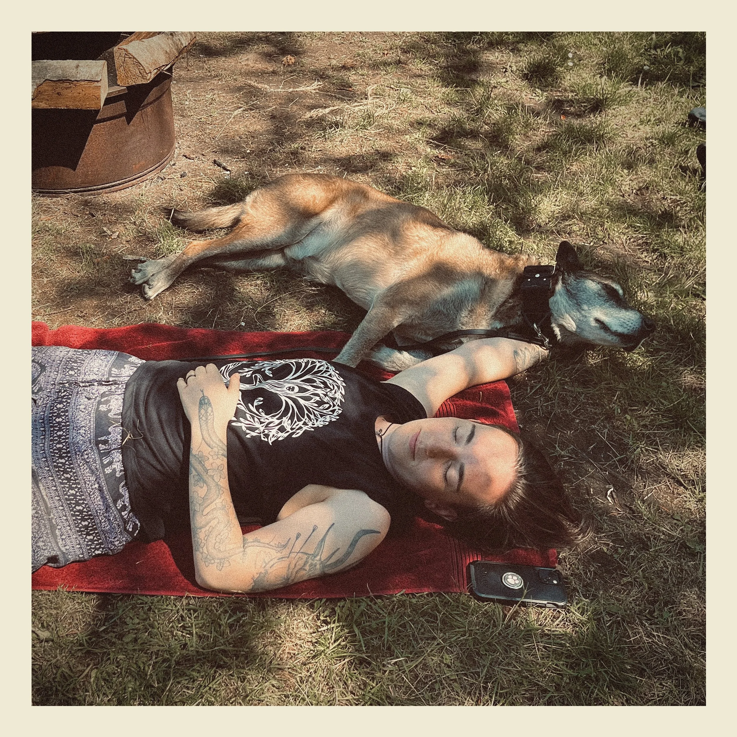 A person with tattoos on their arm lying on the grass with eyes closed, wearing a black shirt with a skull design, red shorts, and a black collar on a dog. A dog with tan and black fur is laying next to them, on its side, on the grass.