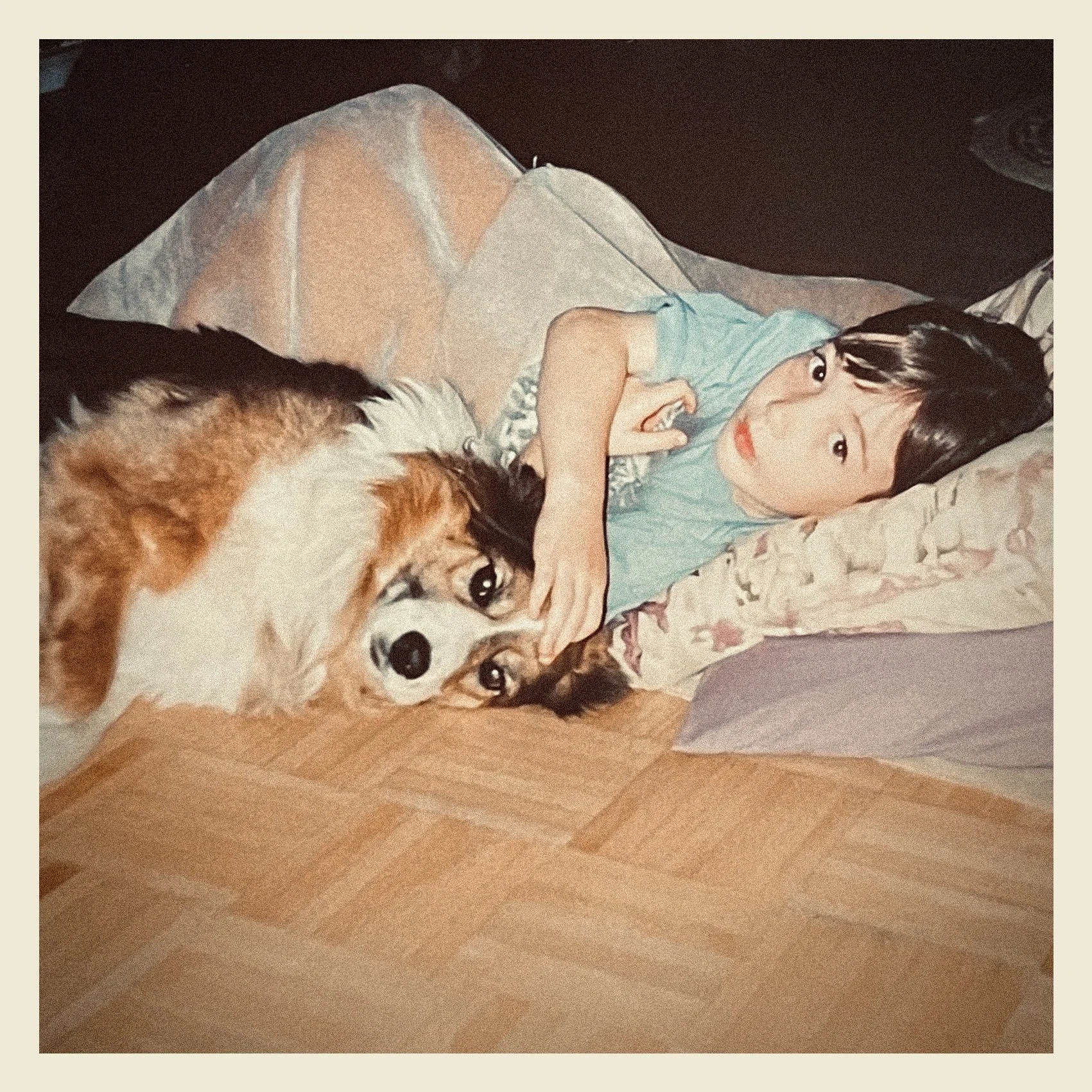 A young girl lying on a bed with a dog, holding the dog's face gently and looking at the camera.