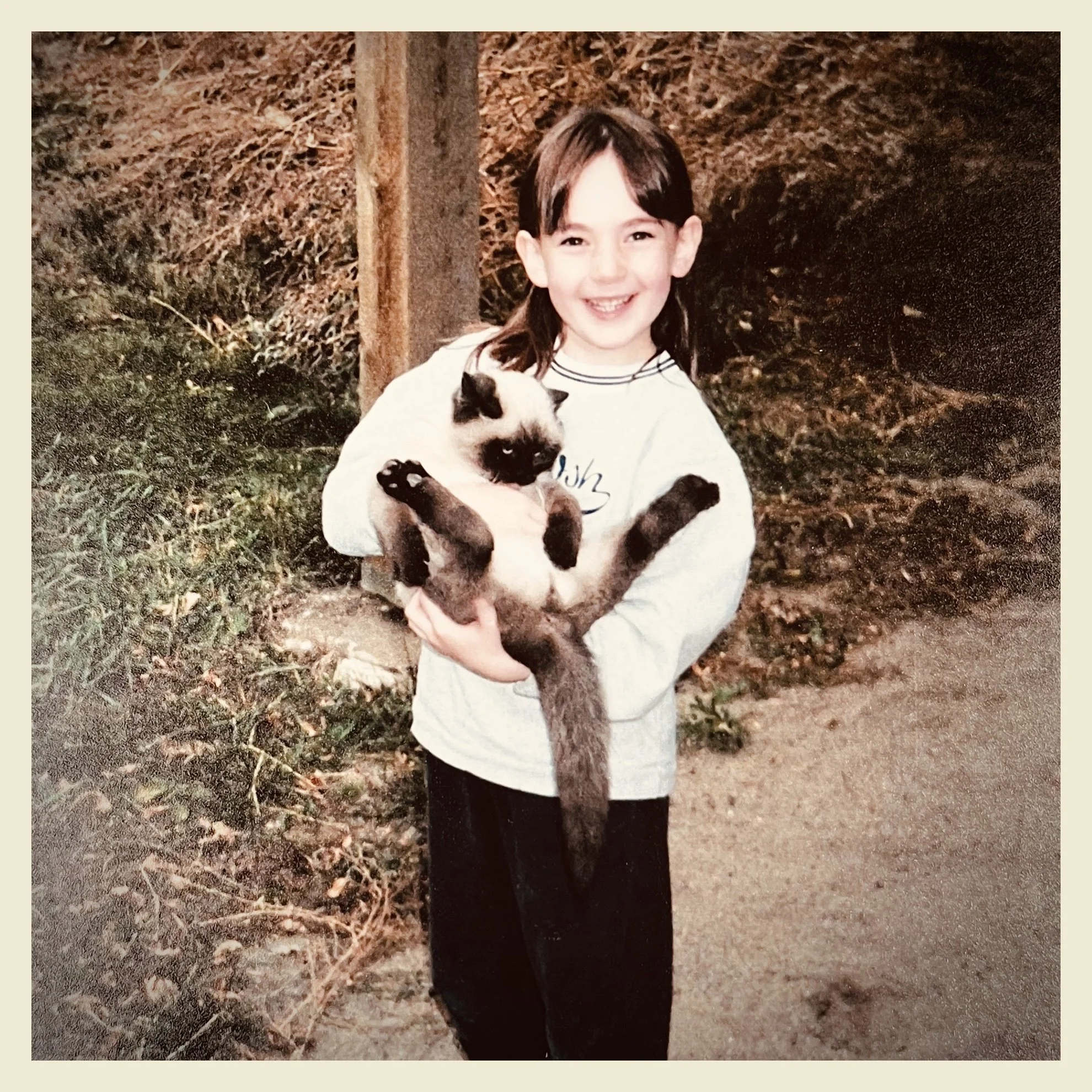 A young girl with dark hair, smiling, holding a small dog with light fur and darker face, in an outdoor setting with trees and leaves.