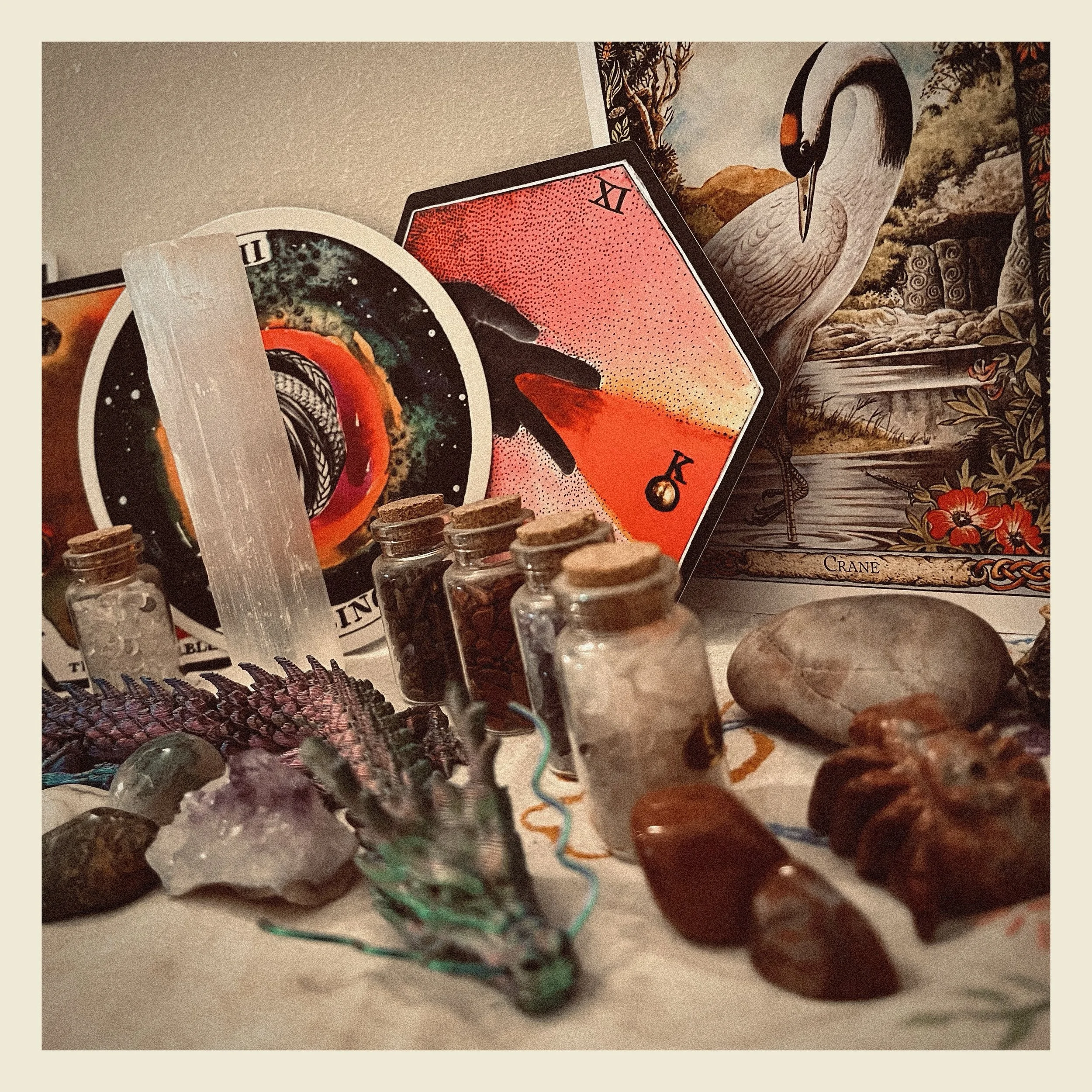 Close-up photo of small glass jars filled with various powders and substances, placed on a shelf. Some jars have cork stoppers, and there is a colorful woven item nearby. In the background, there are tarot cards, including one with a swan illustration and a landscape. The scene appears to be a collection of mystical or spiritual items.
