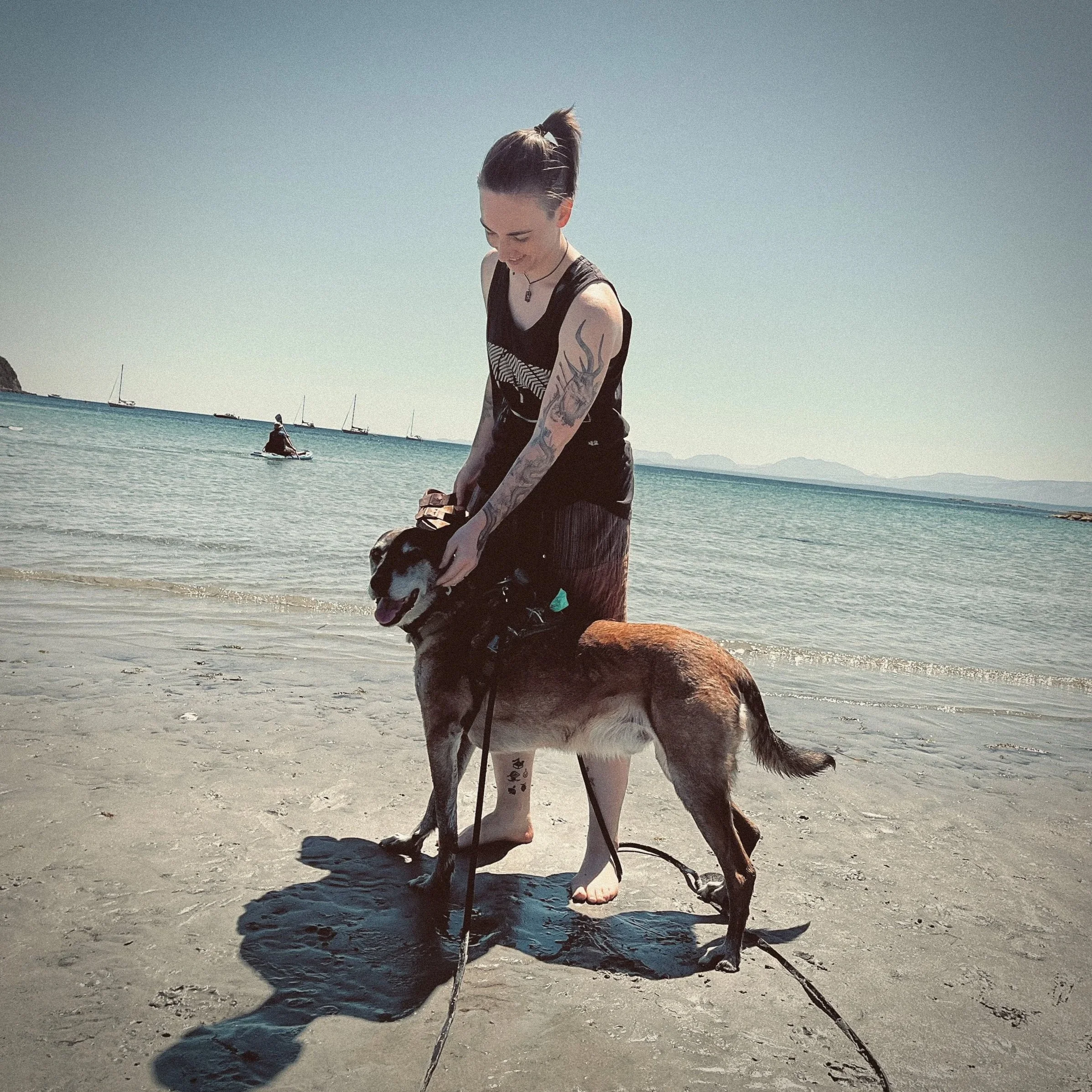 A young woman with tattoos on her arms and a ponytail is standing on the beach with a dog, near the water, with sailboats in the background on a clear day.