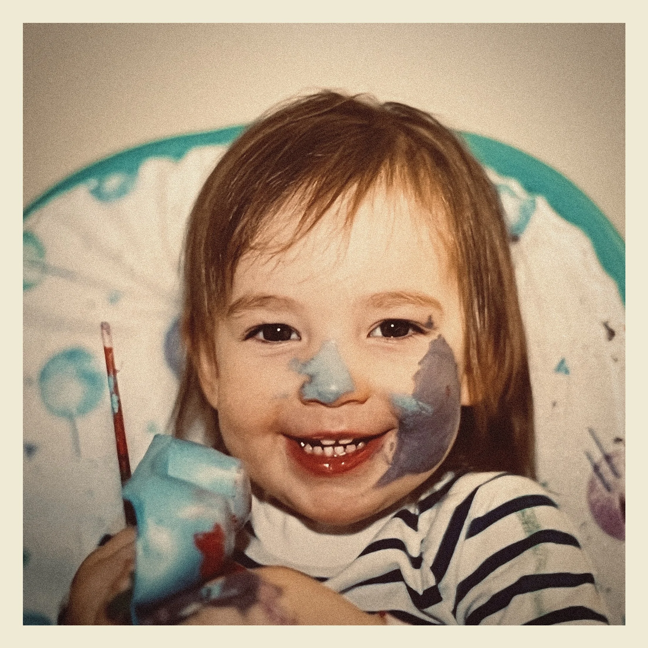 Close-up of a smiling baby with brown hair, wearing a black and white striped outfit, lying on a white surface with a blue patterned blanket nearby.