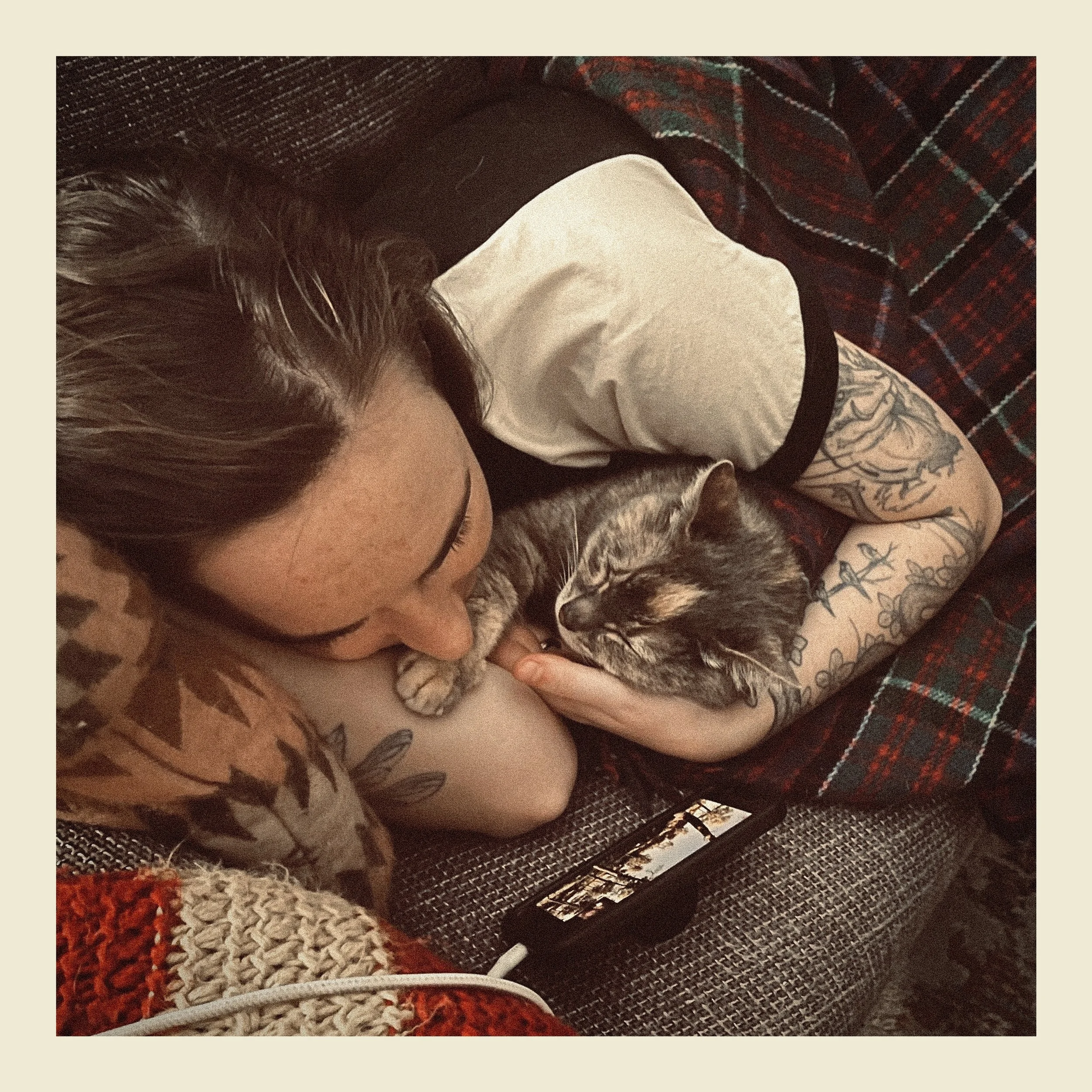 Person with tattoos cuddling a small cat on a brown and red patterned carpet.