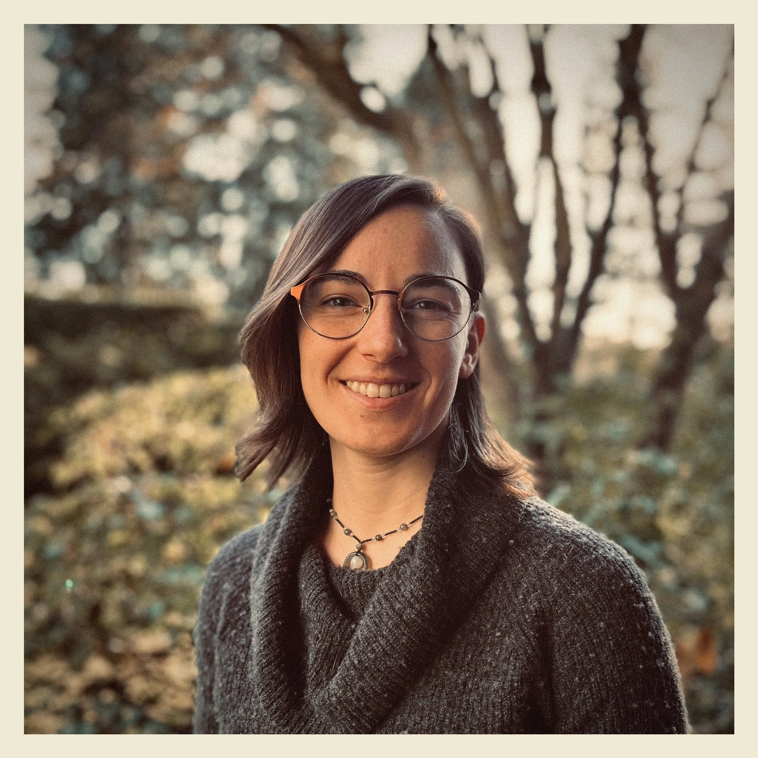 A woman with shoulder-length brown hair, wearing glasses and a black textured coat, smiling outdoors with a tree in the background during autumn.
