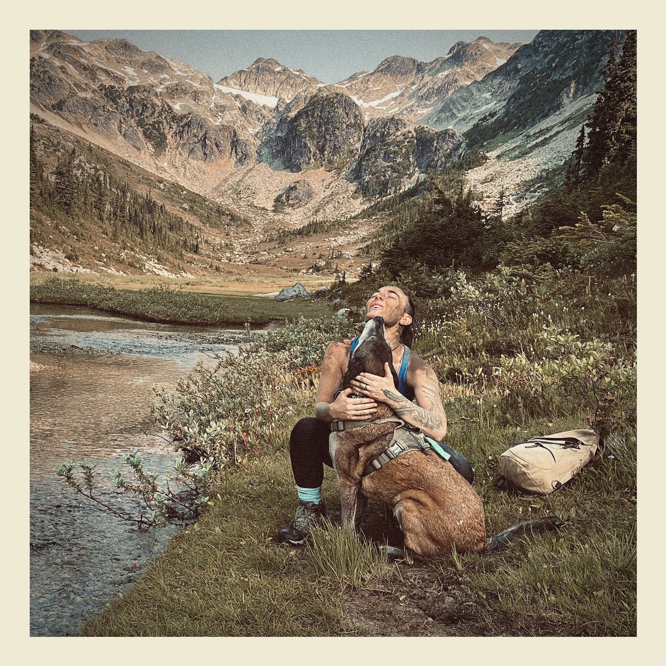 A person with tattoos and long hair is hugging a dog in a scenic mountain landscape with rocky peaks, a river, and green vegetation.