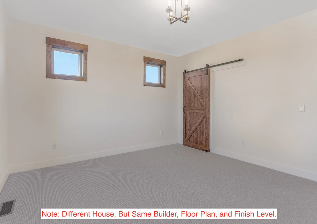 Empty room with beige walls, two small square windows with wooden trims, a sliding barn door, a modern ceiling light fixture, and a light gray carpeted floor.