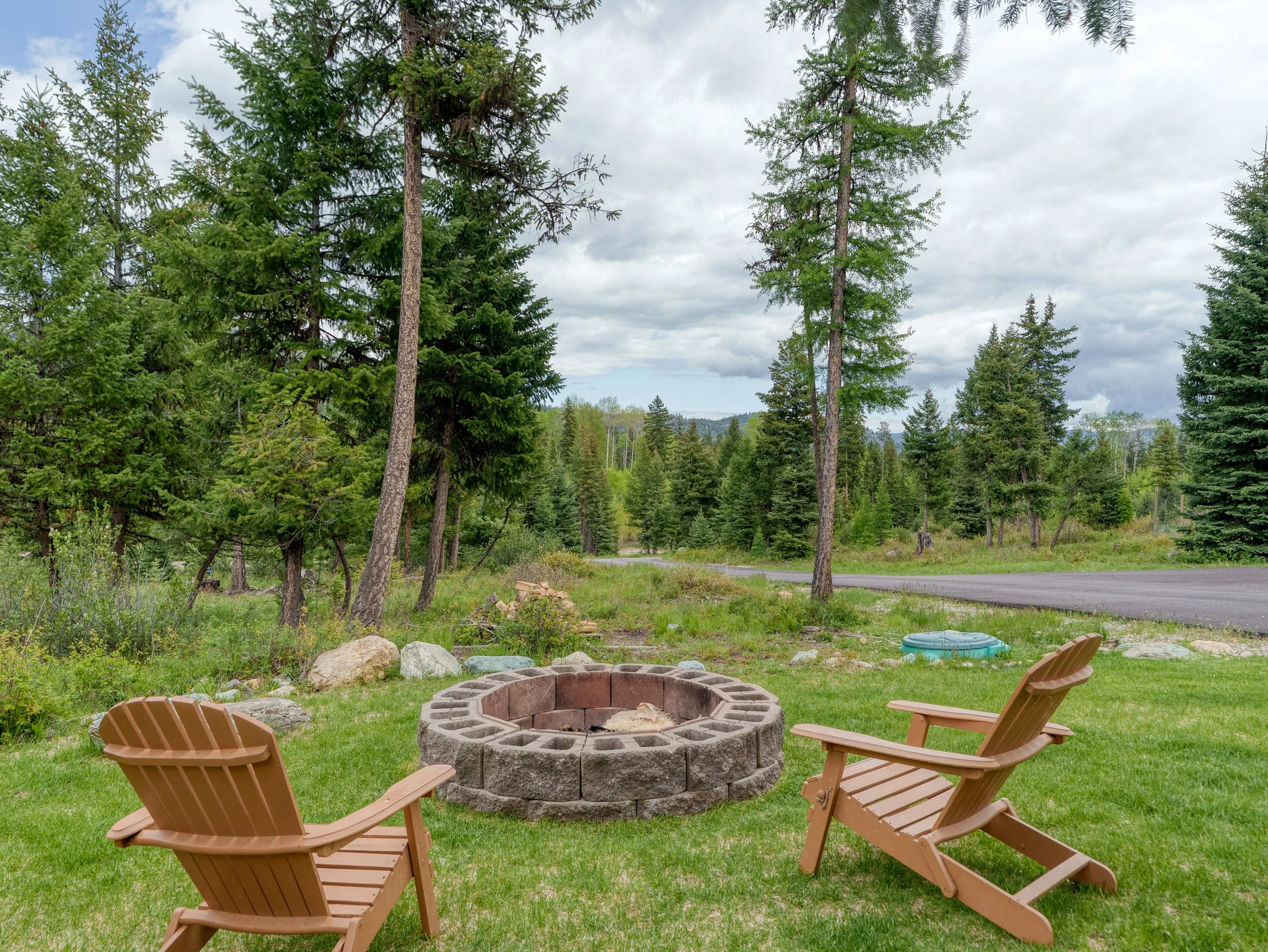 View of a backyard with two wooden chairs around a circular fire pit, surrounded by grass and rocks, with tall pine trees and a cloudy sky in the background.
