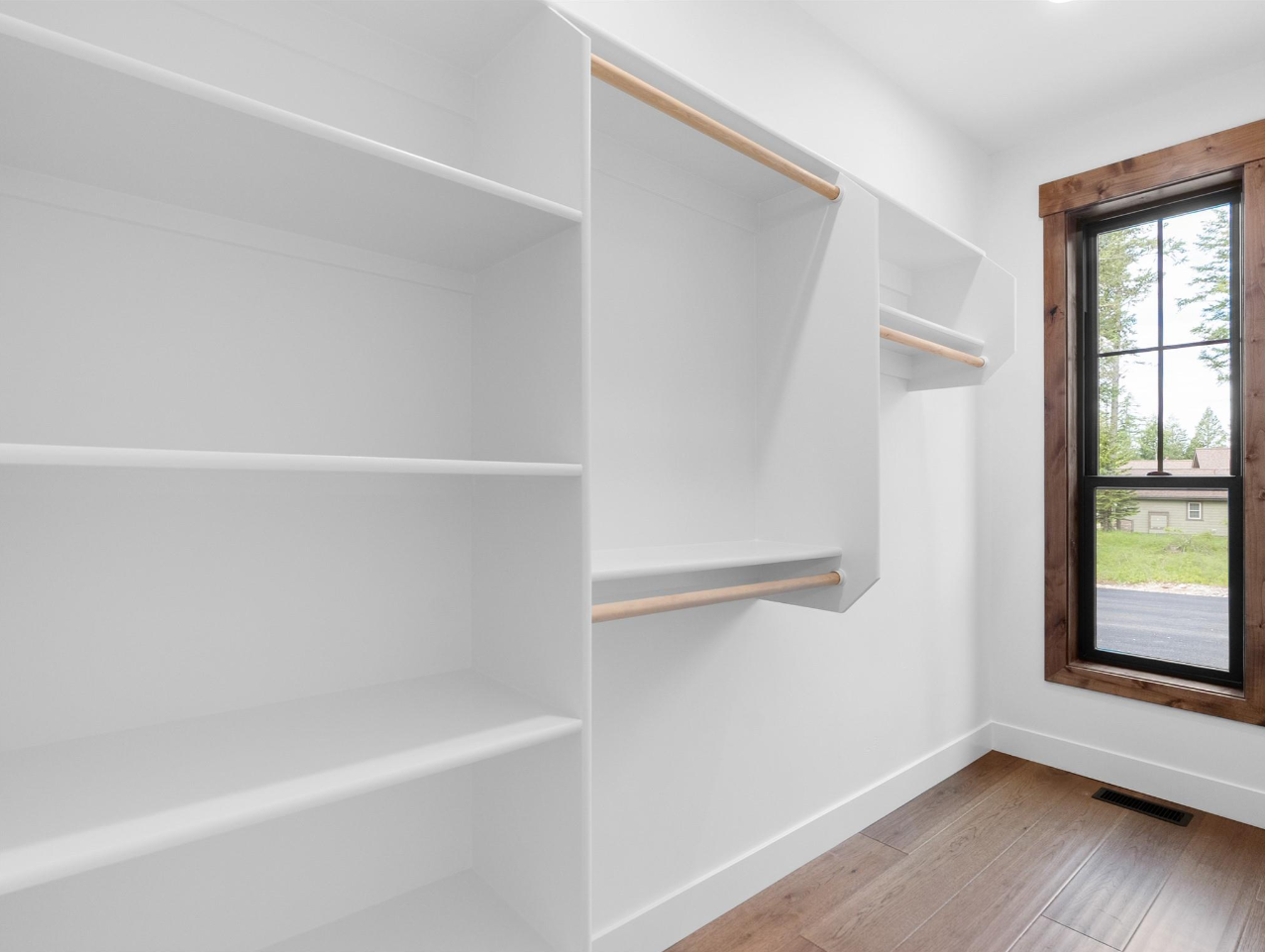 Empty walk-in closet with white shelves and rods, wooden trim around window, hardwood floor, and a floor vent.