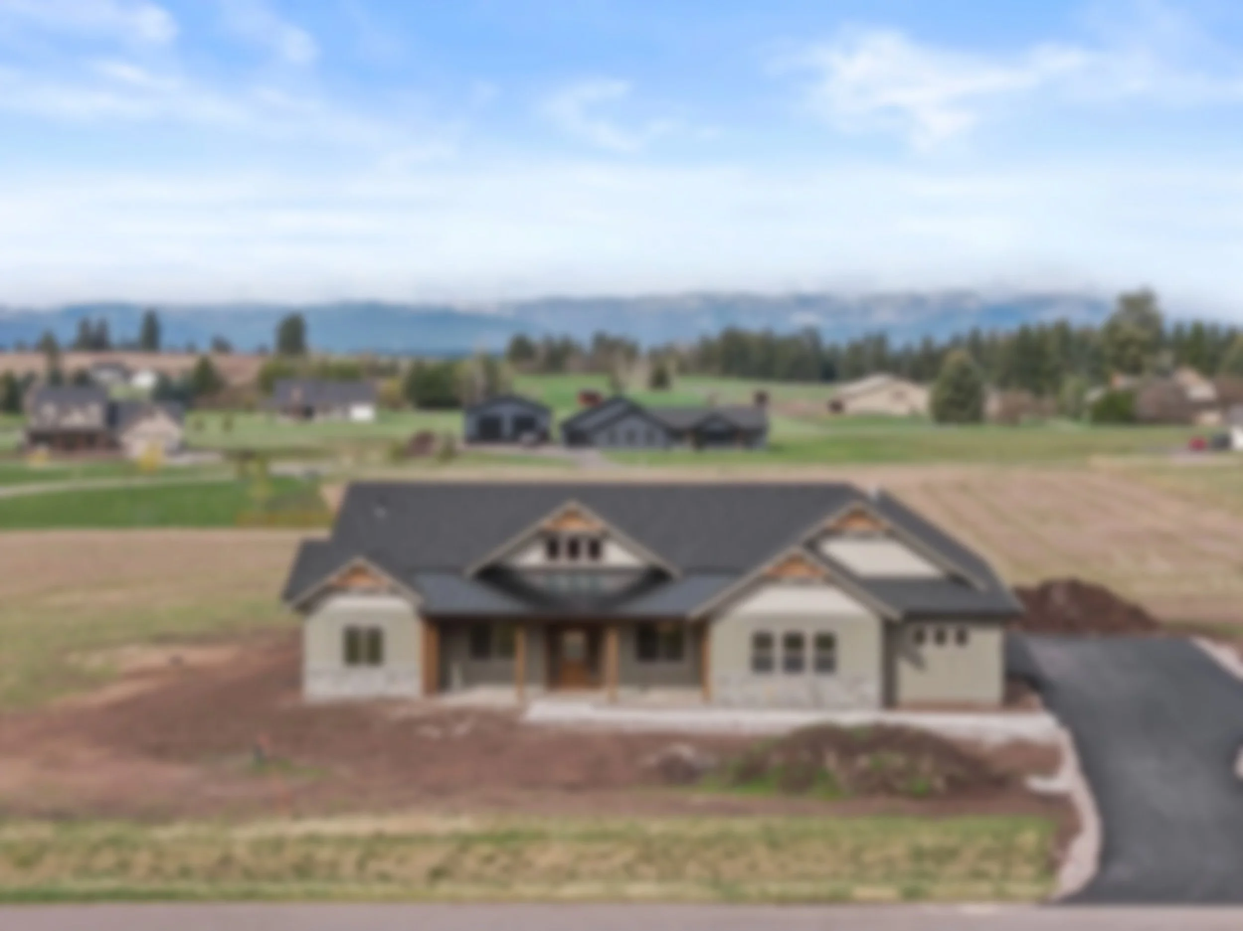 Newly built house with gray siding, stone foundation, large front windows, and black roof, located in a suburban area with other houses and open fields, mountains in the background under a partly cloudy sky.
