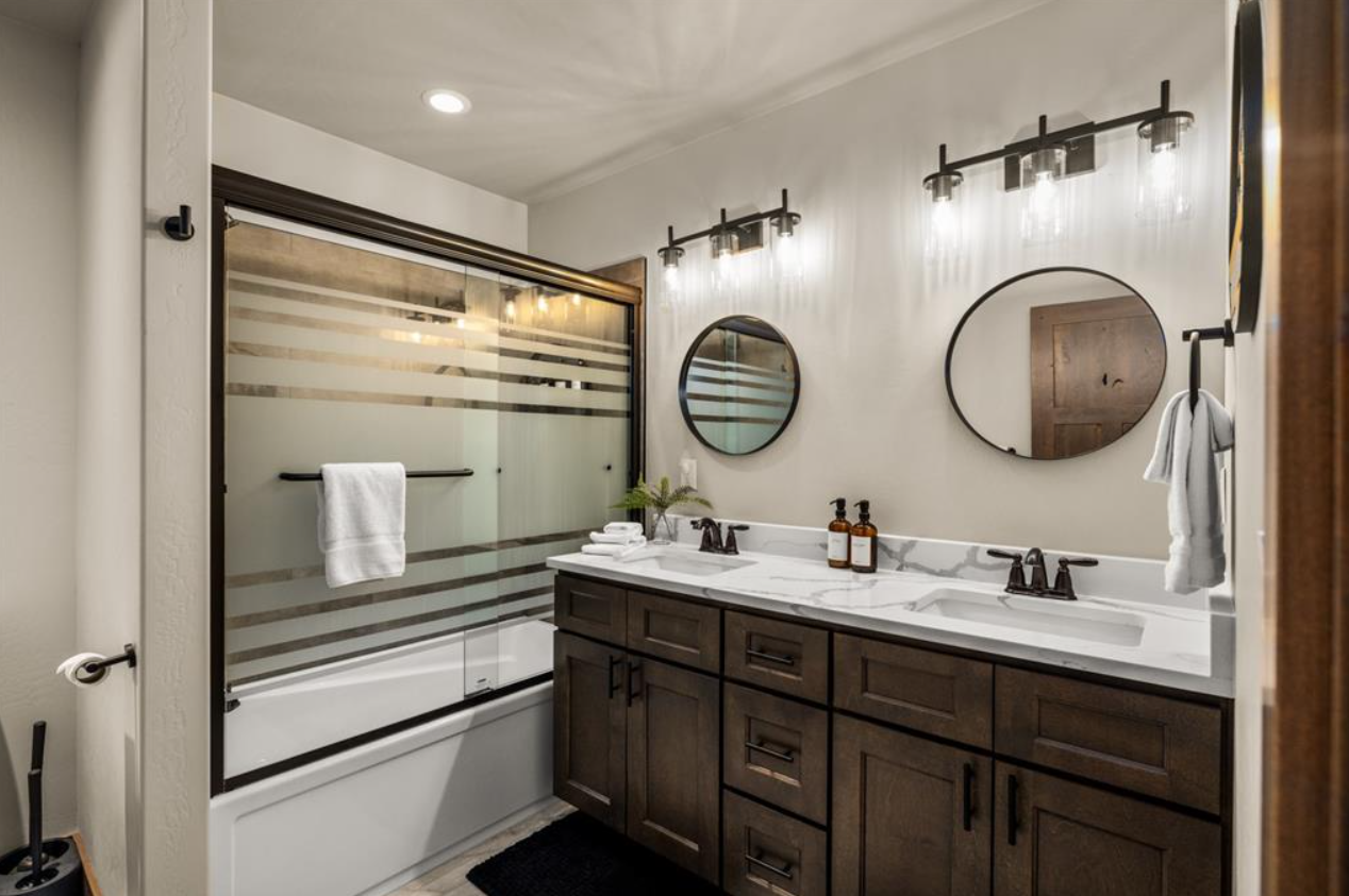 A modern bathroom with a double vanity, dark wood cabinets, and a white countertop. Two round mirrors hang above the sinks, and there are two wall-mounted light fixtures. The bathtub with a glass sliding door has horizontal frosted stripes, and a whi