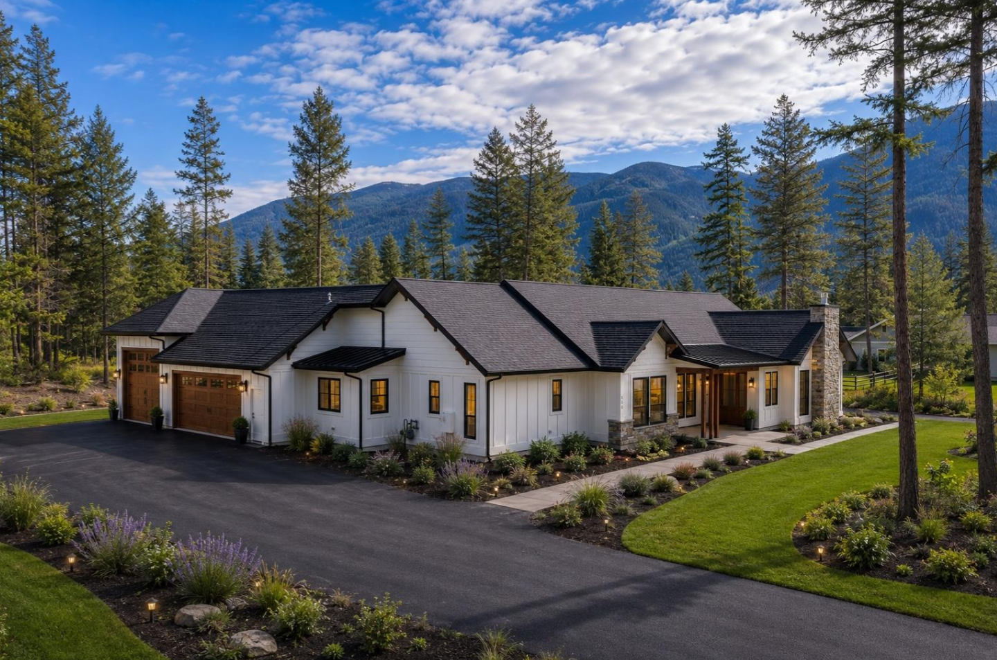 Modern white house with black accents, stone chimney, and attached garage surrounded by well-maintained lawn and garden, set against a backdrop of tall pine trees and mountain range.