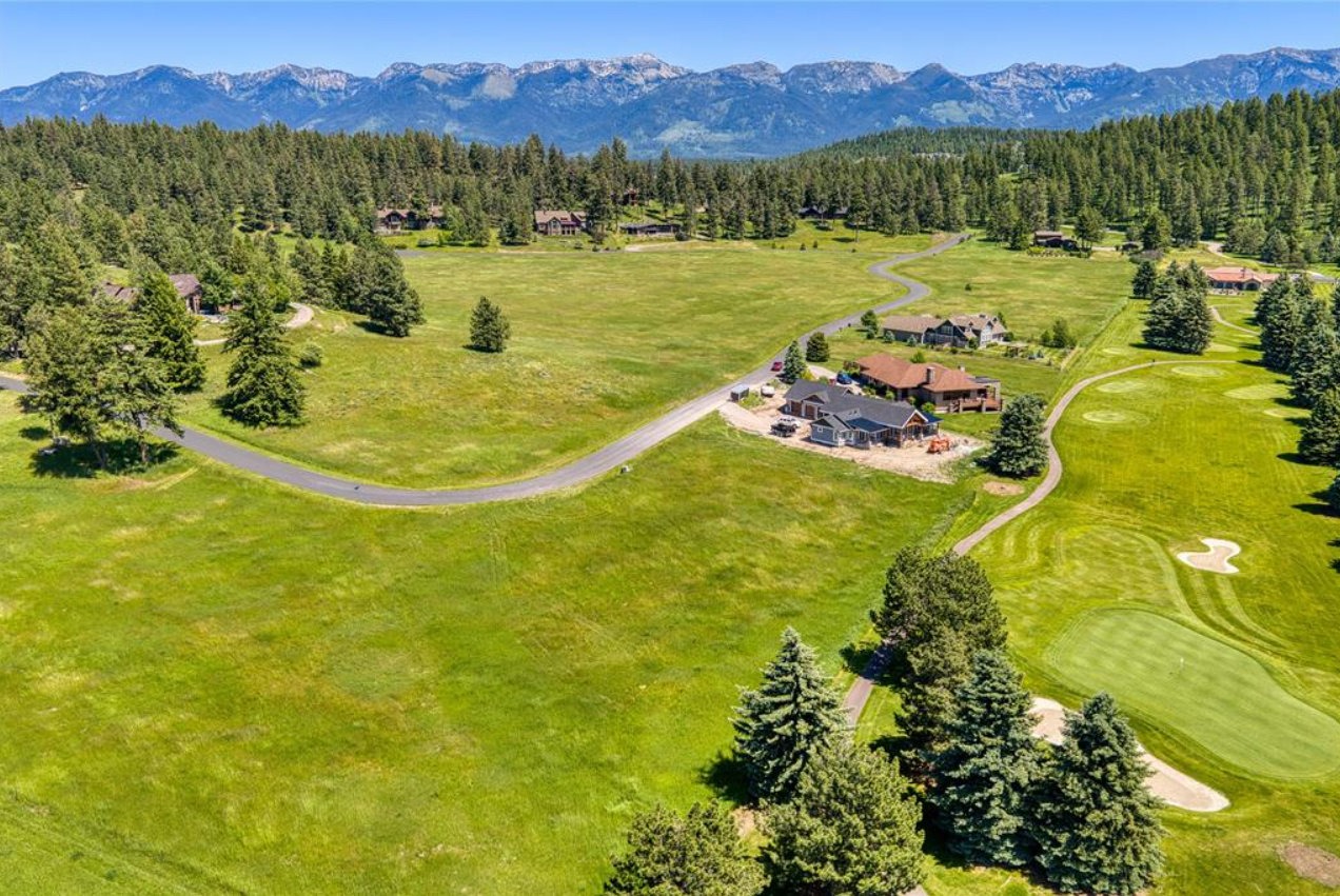 Aerial view of a golf course across green fields with trees, houses, and mountains in the background.
