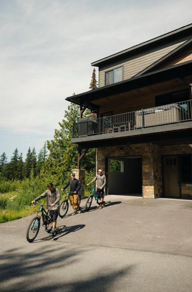 Three men riding bikes near a house with a stone and wood exterior, overlooking a green landscape with trees.