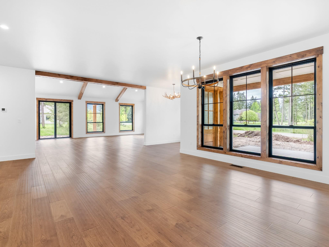 Empty room with large windows, wood flooring, white walls, and wooden beams, with outdoor greenery visible through the windows.