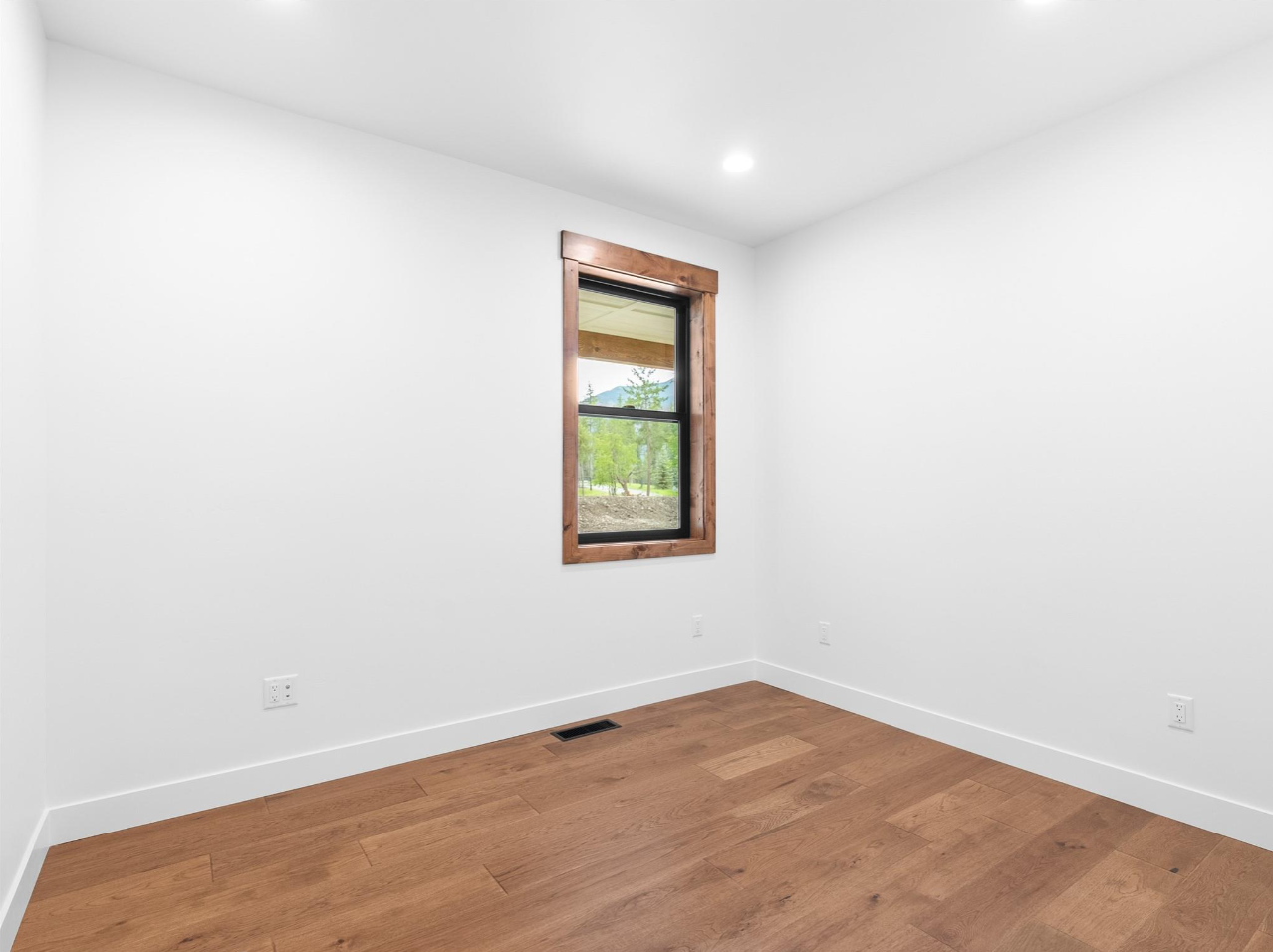 Empty room with white walls, wooden floor, a window with wooden trim showing outdoor trees and a mountain, and electrical outlets.