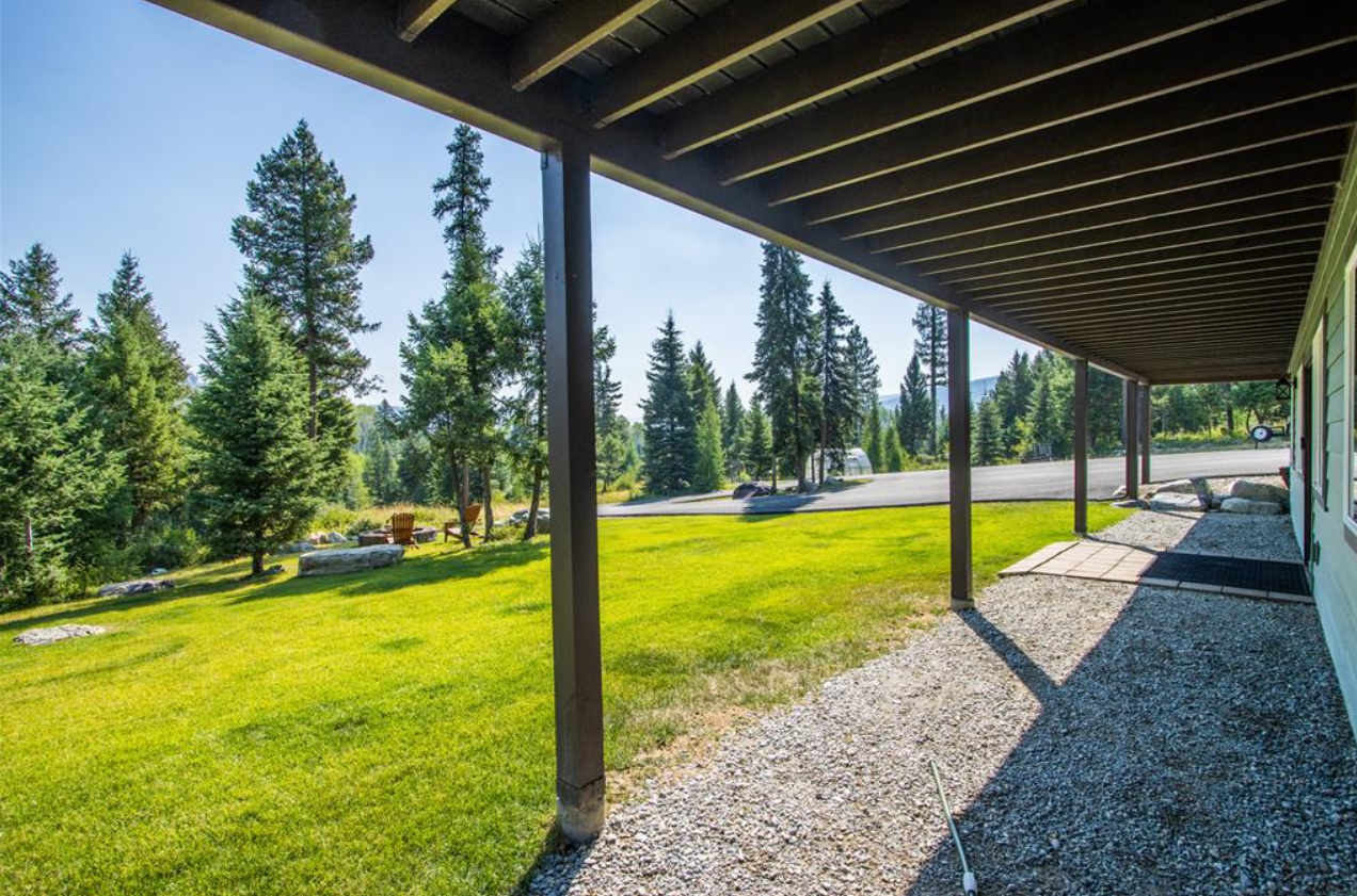 View from a porch looking out onto a grassy yard with trees and a mountain in the distance. The porch is supported by wooden posts and has a gravel walkway.