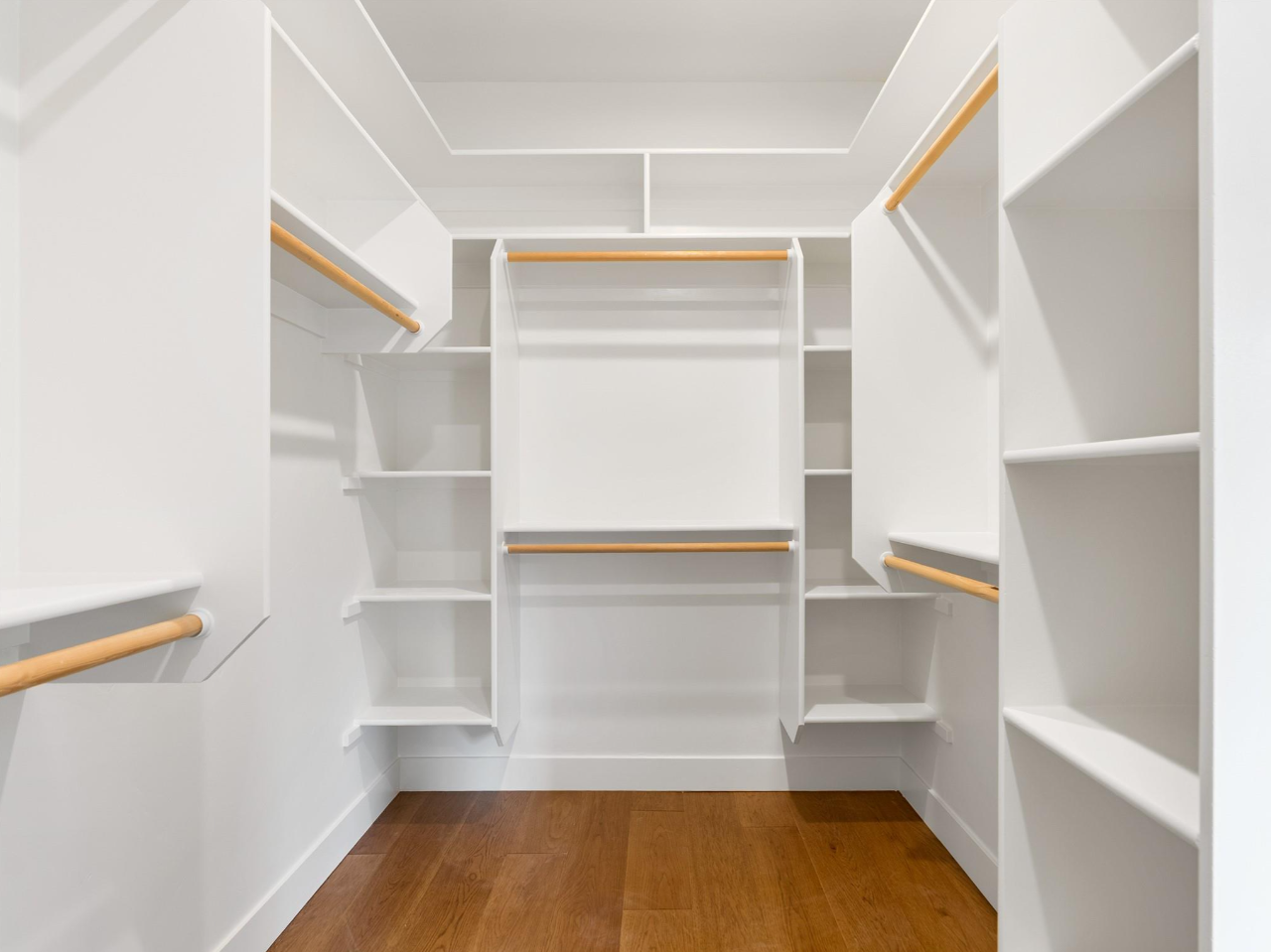 Empty white walk-in closet with wooden shelves and hanging rods, hardwood floor.
