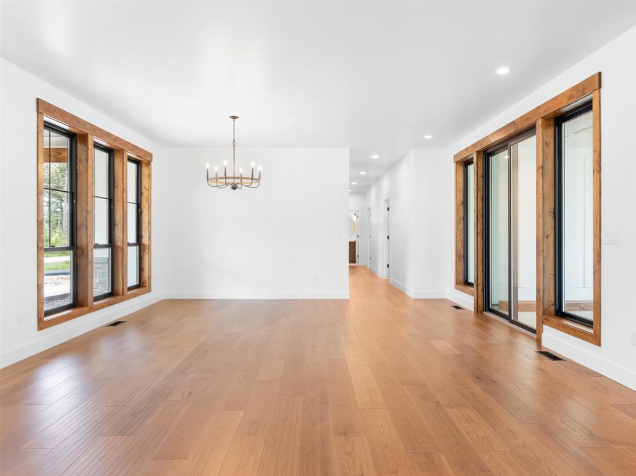 Empty living room with wooden floors, white walls, large windows with wooden frames, and a chandelier.