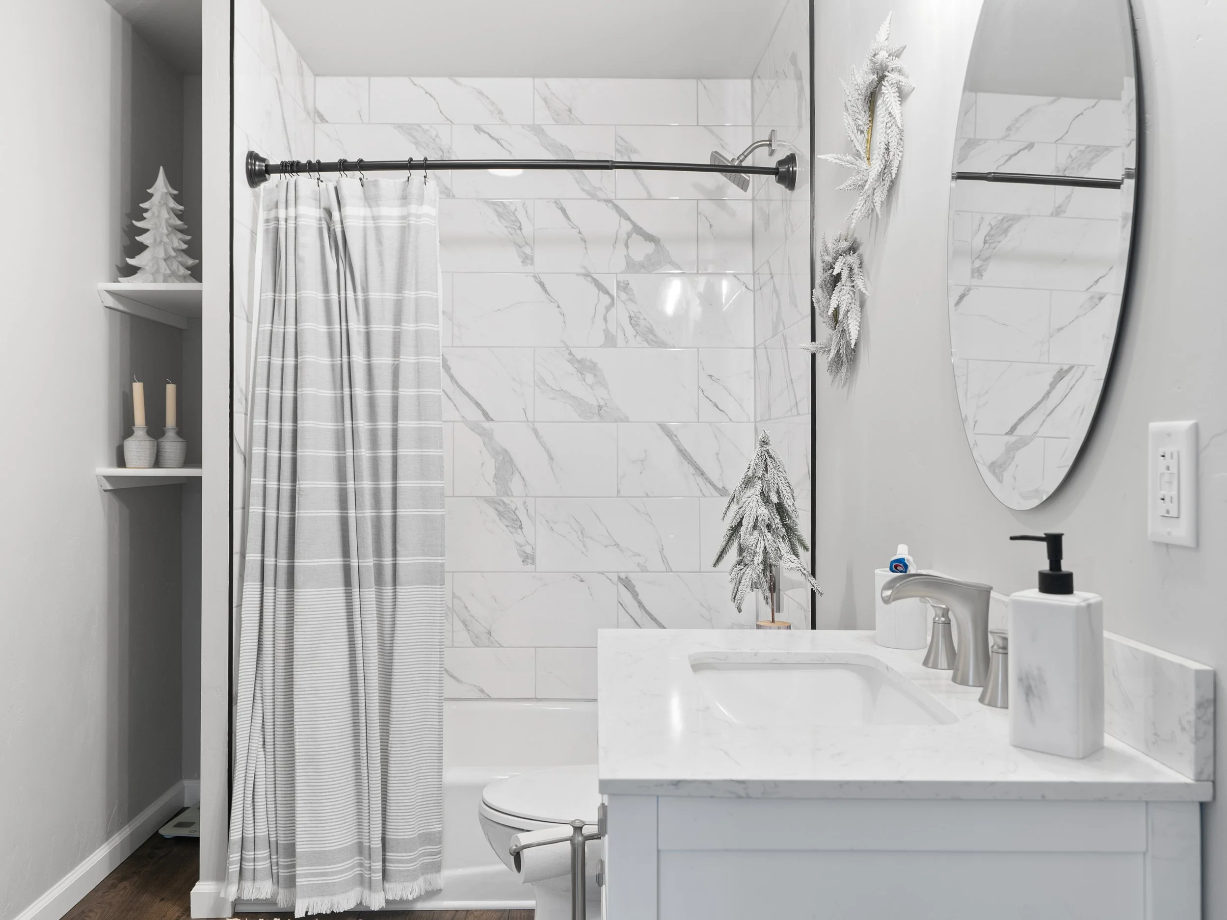 A modern bathroom with white marble tiles on the wall and a white vanity with a white marble countertop. There is a large oval mirror on the right with a decorative wreath hanging beside it. A small potted plant sits on the vanity, and a black faucet