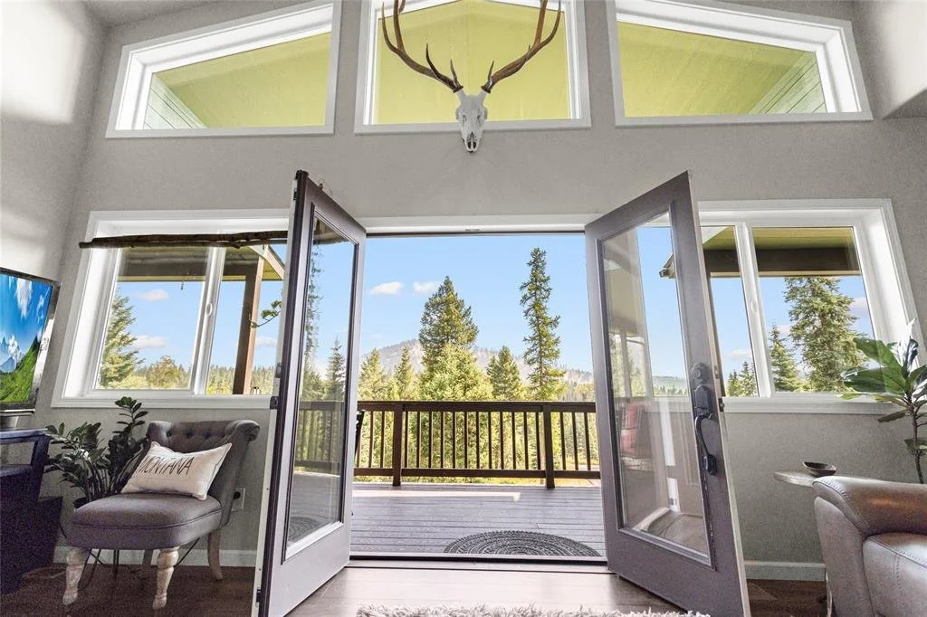 Living room with large open glass doors leading to a deck overlooking a forested landscape, a mounted deer skull above the doors, and windows allowing natural light in.