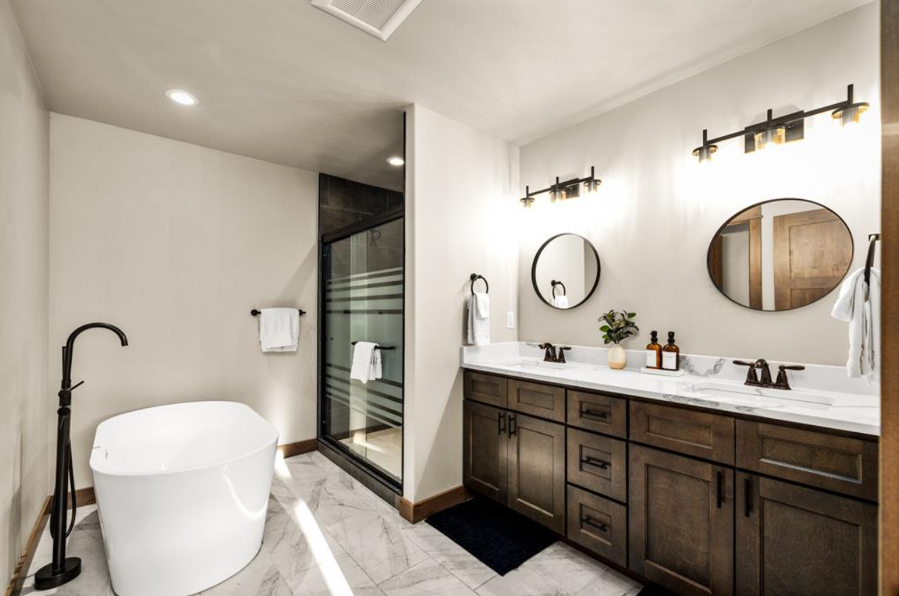 Modern bathroom with a white freestanding tub, double vanity with dark wood cabinets, marble countertop, oval mirrors, and glass shower enclosure.