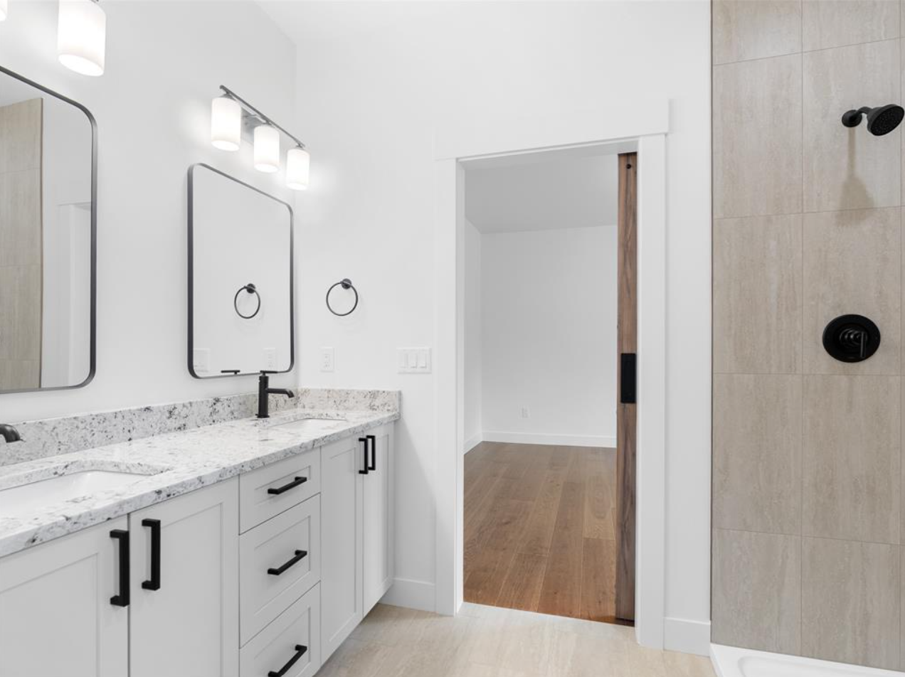 Modern bathroom vanity with white cabinets and black handles, granite countertop, two mirrors, black faucets, and a wall-mounted light fixture. Adjacent walk-in shower with beige tiles and black fixtures.