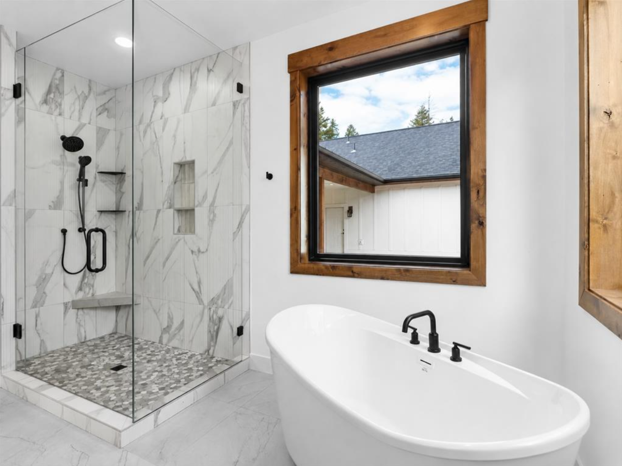 Bathroom with walk-in shower with marble tiles, a freestanding bathtub, and a large window with wood trim showing an outdoor view.