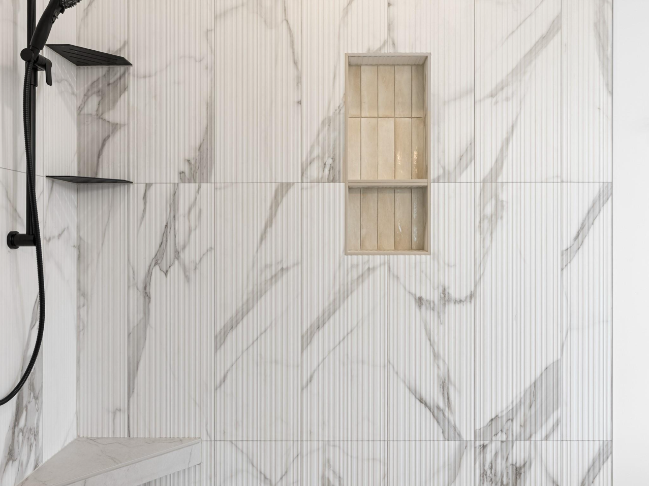 Modern shower with white marble tiles featuring gray veining, a black showerhead, two small black shelves, and a built-in beige niche.