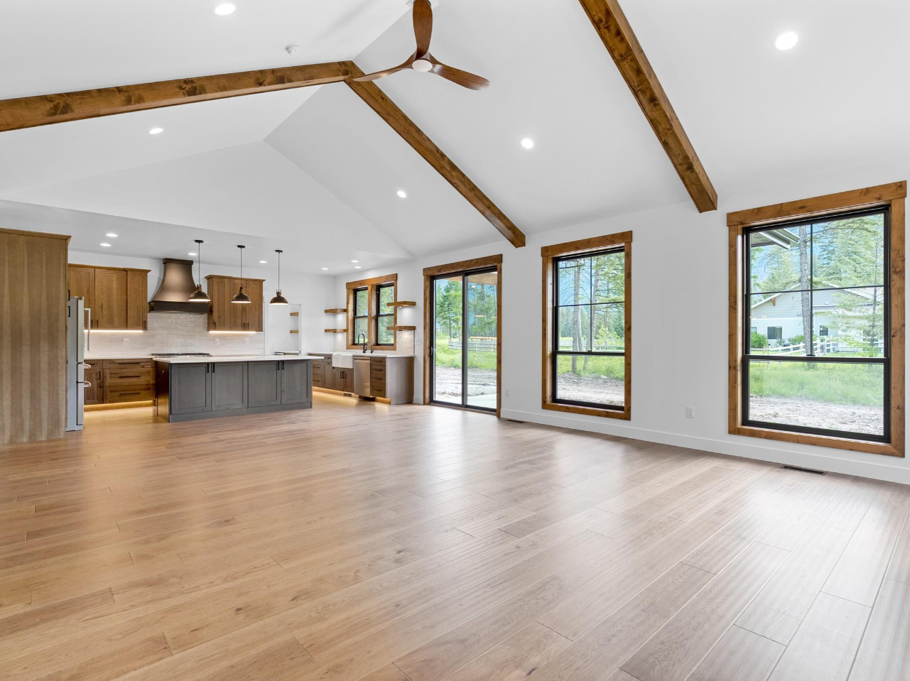 Bright open-concept living area with large windows, wooden beams, and a kitchen with wooden cabinets and a gray island.