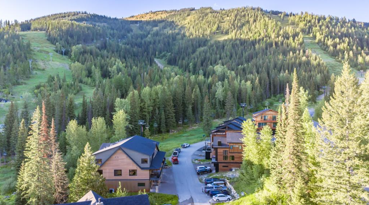Mountain resort with chalet-style buildings, surrounded by lush green forest, ski slopes, and mountain peaks in the background.