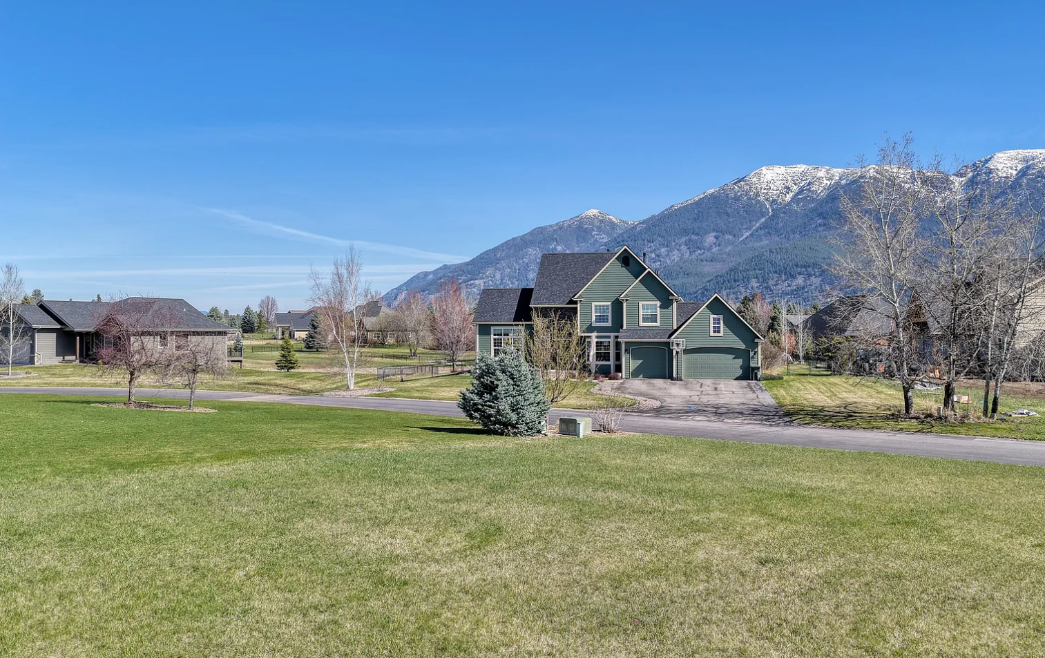 A suburban neighborhood with a green lawn, trees, and houses at the base of snow-capped mountains under a clear blue sky.