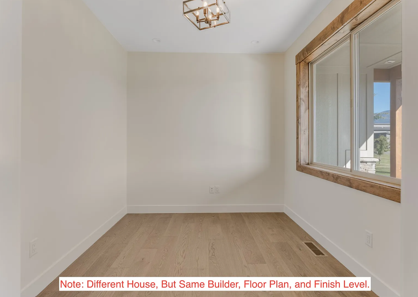 Empty room with white walls, light wood flooring, a large window with wood trim, and a modern ceiling light fixture.