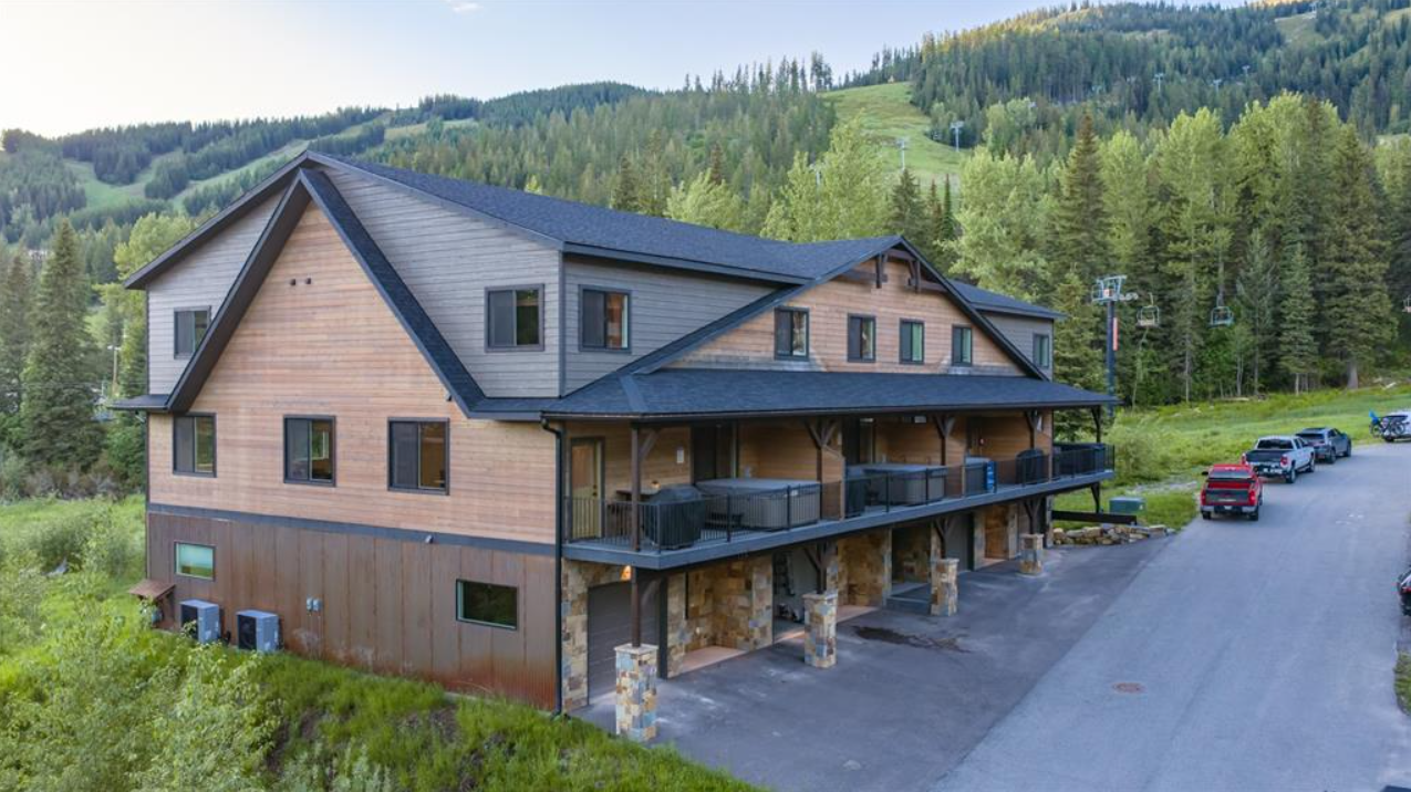 A multi-story wooden mountain lodge with a black roof, surrounded by a forested hillside and a ski slope, with parked cars nearby.