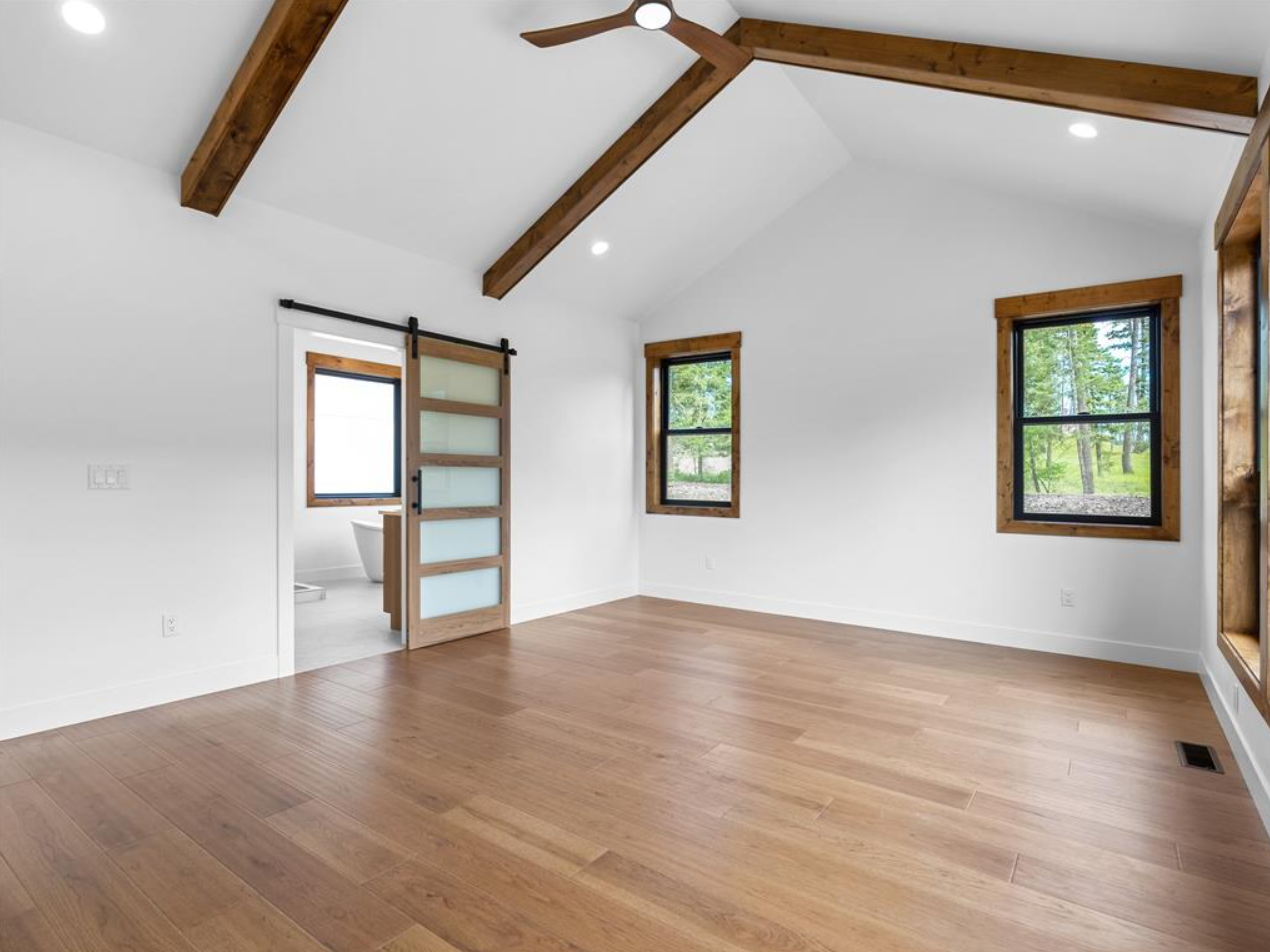 Empty room with wooden floors, white walls, and exposed wooden beams on the ceiling. There are three windows with wooden frames and a sliding door leading to a bathroom.