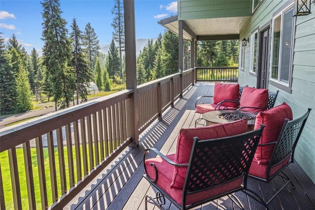 Balcony with four red cushioned chairs and a small round fire pit table, overlooking a lush, green forested area with tall trees under a partly cloudy sky.