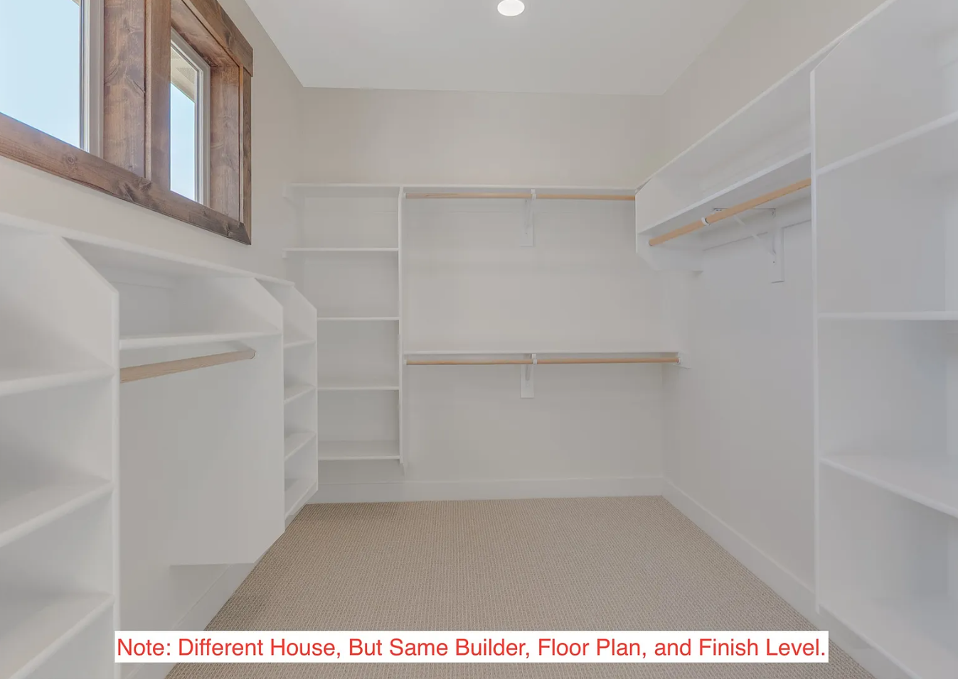 Empty walk-in closet with built-in white shelving, wooden rods for hanging clothes, a small window with a wooden frame, and beige carpeted floor.