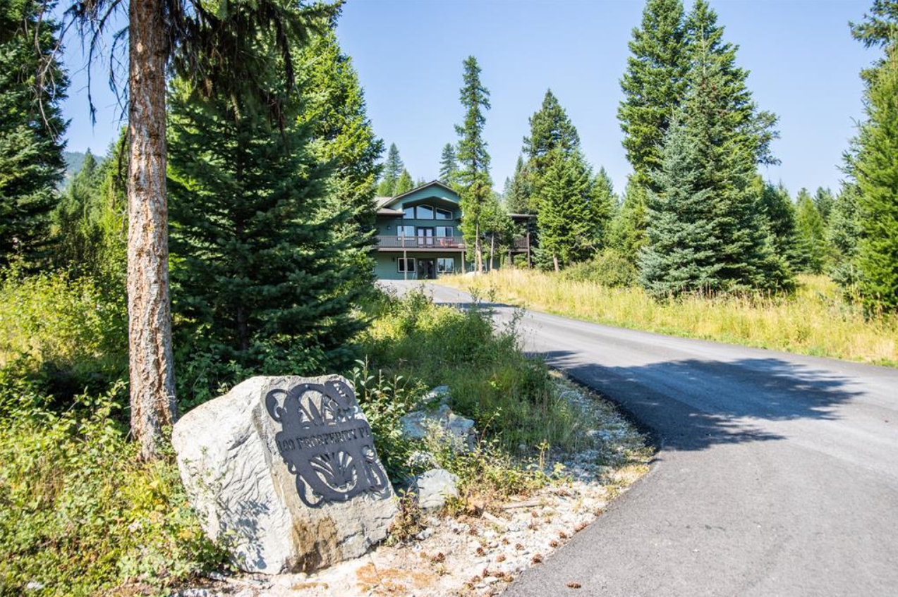 A gravel driveway curves past a large rock with a metal sign that reads "No Property" and a decorative pattern. There are tall pine trees and a house with a balcony visible in the background, surrounded by lush green forest on a sunny day.