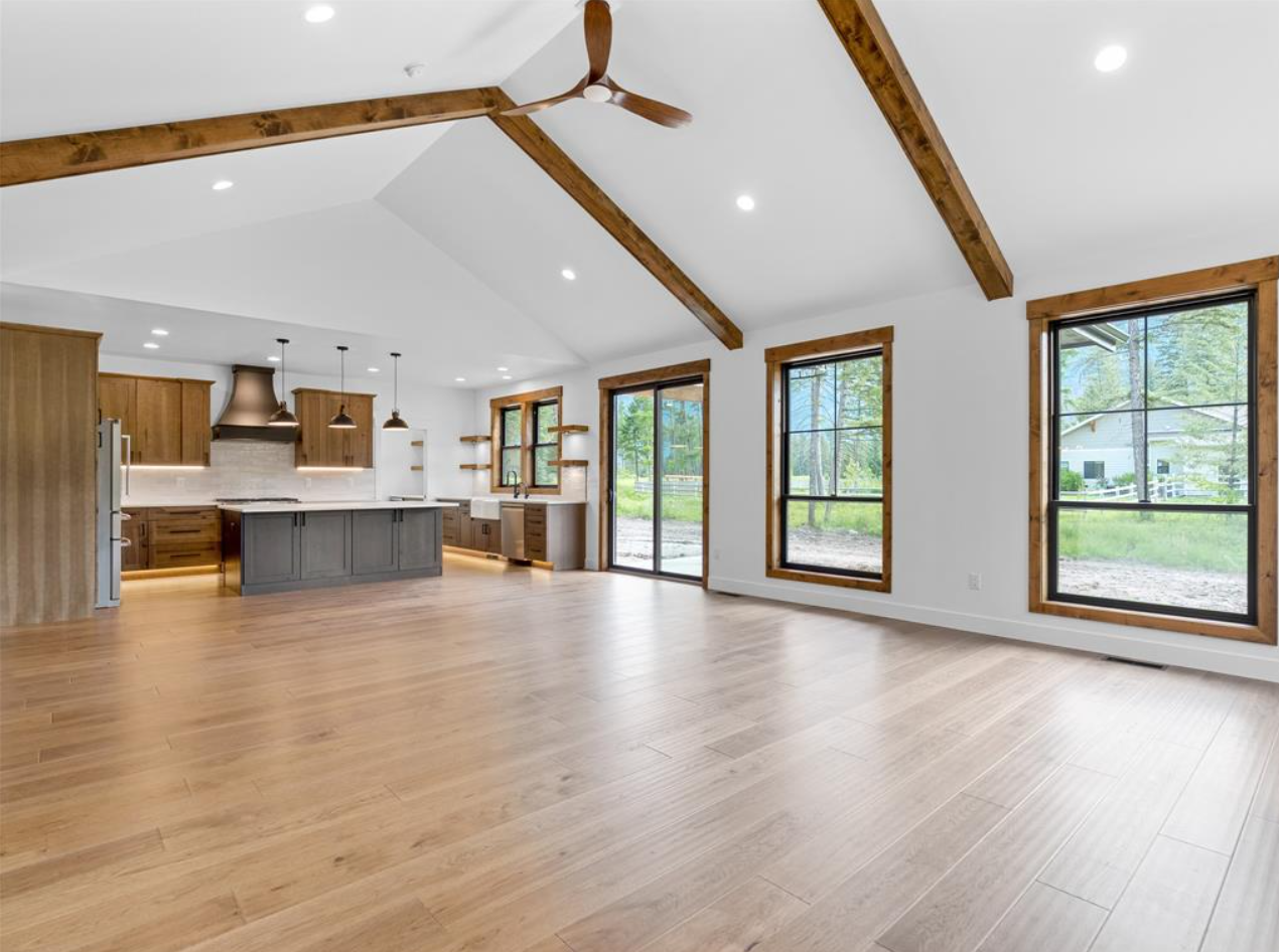 Open-concept living space with a kitchen and large windows, featuring wood flooring, white walls, wooden beams on the ceiling, and a ceiling fan.