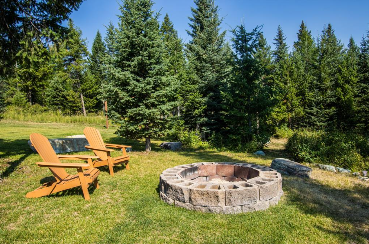 Two wooden Adirondack chairs beside a stone fire pit on a grassy lawn, with a backdrop of tall green pine trees and a clear blue sky.