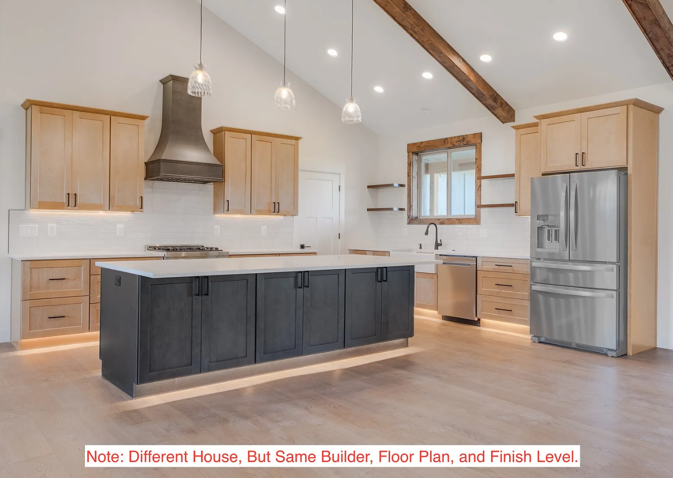 Modern kitchen with light wood cabinets, a black kitchen island, stainless steel refrigerator, and a range hood with pendant lights above.