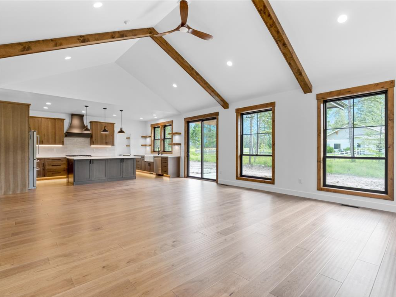 Open-concept living and kitchen area with hardwood floors, large windows, white walls, wooden ceiling beams, and modern kitchen cabinets and island.